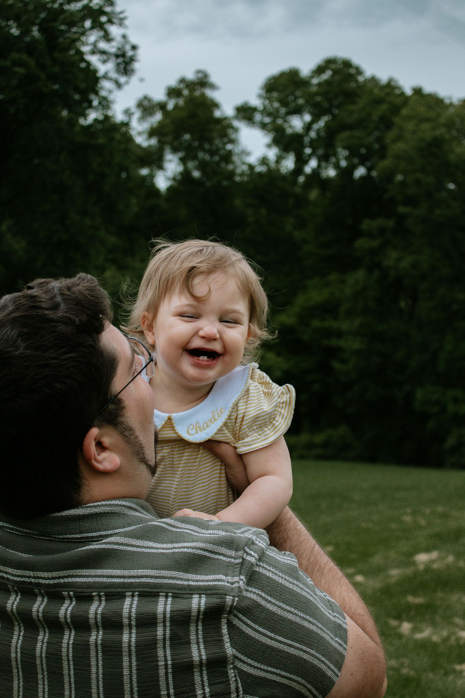 A laughing baby girl in a yellow dress and white bib is held up by a man in a striped shirt outdoors at French Park in Cincinnati, Ohio