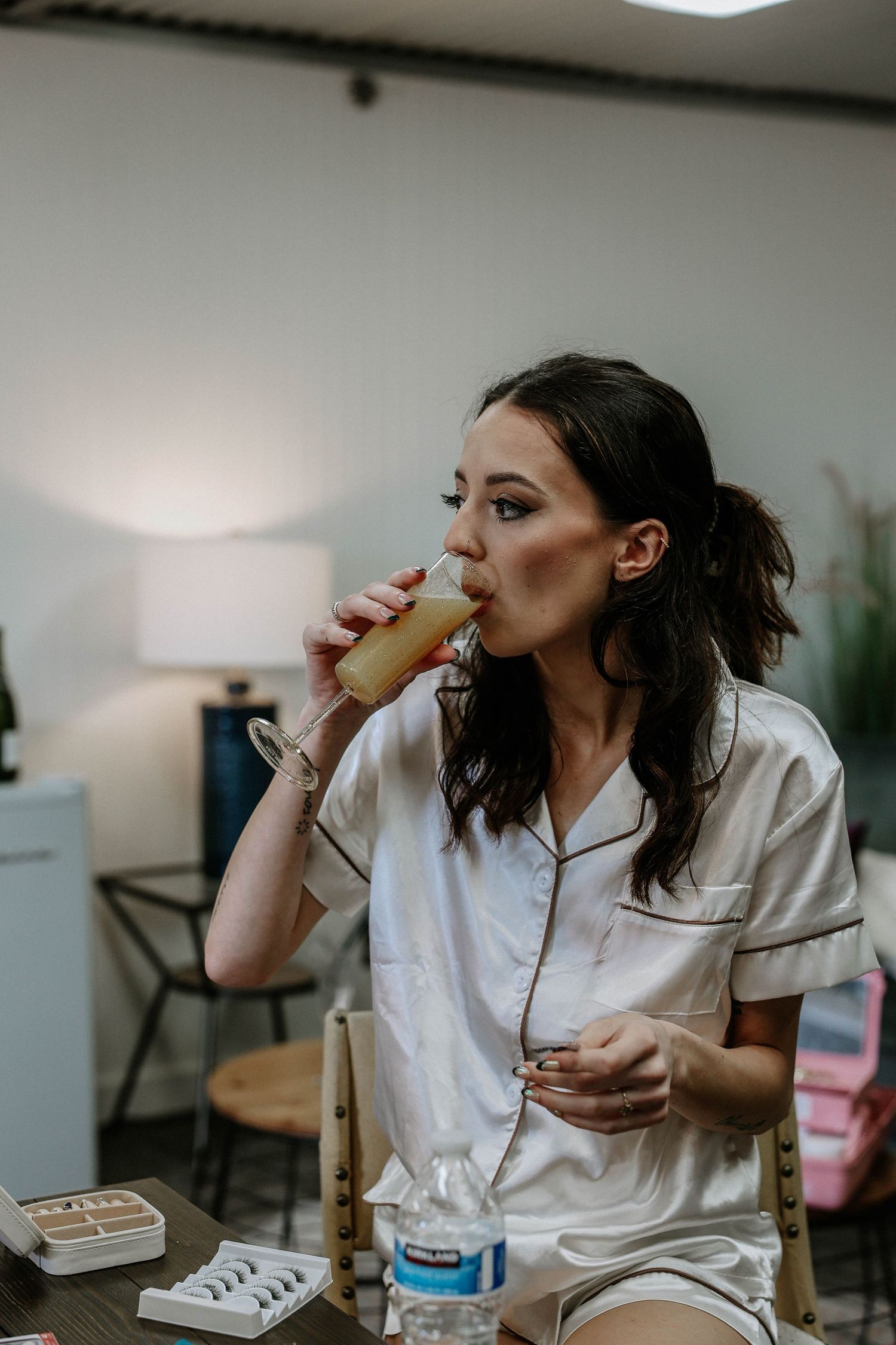 Bride in white satin pajamas sipping a drink at a vanity table with false lashes and water bottle nearby.