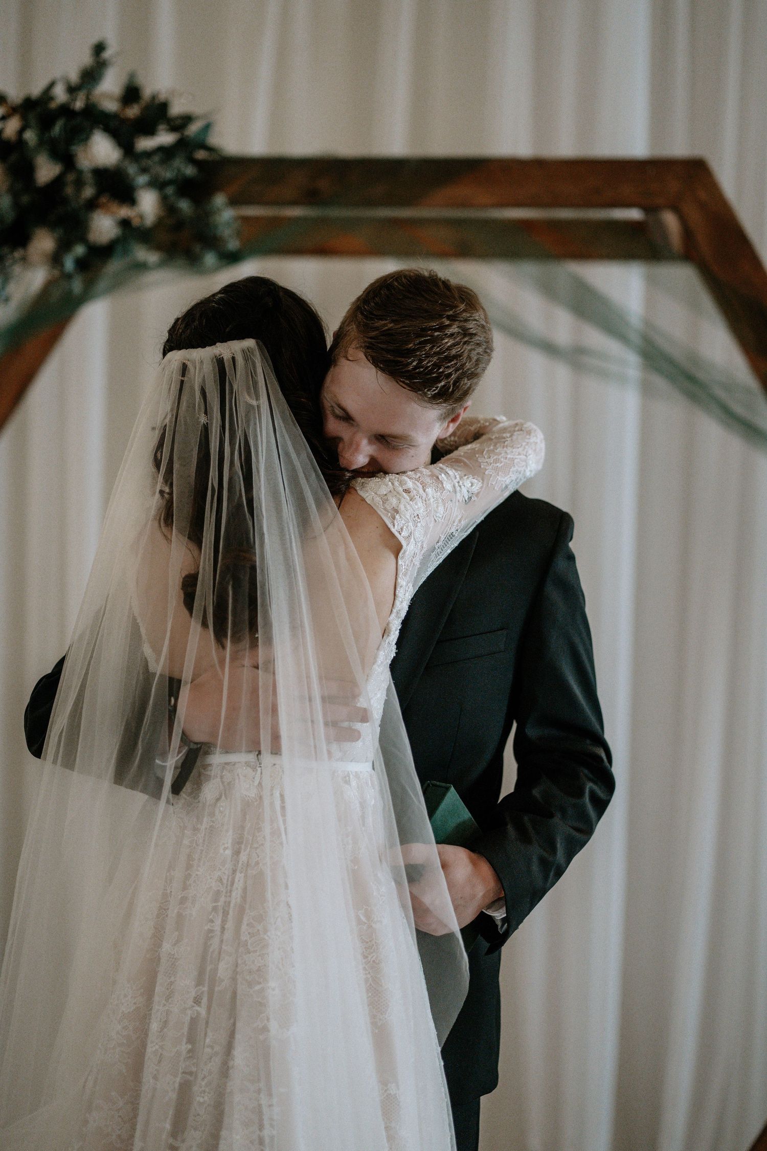 Bride and groom share an emotional embrace at the altar beneath a wooden hexagon arch with floral decor.
