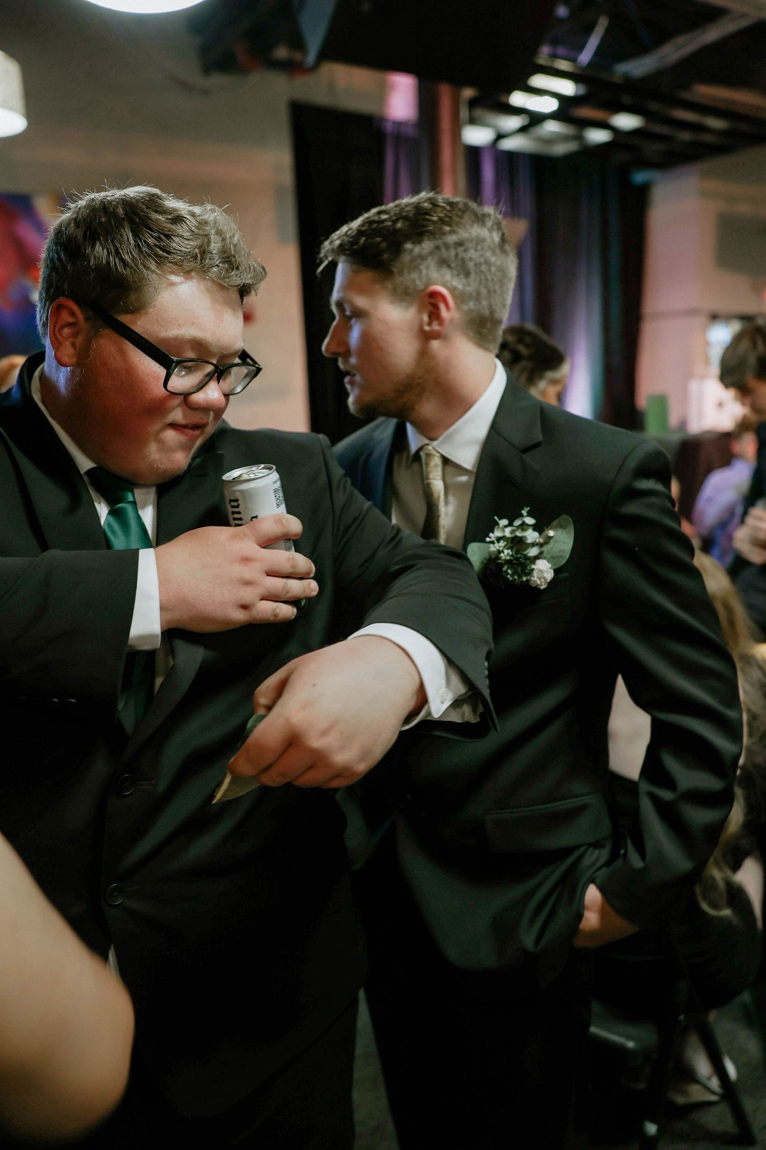 Two young men in black suits interact at a formal event, one wearing glasses and a teal tie, the other with a boutonniere.