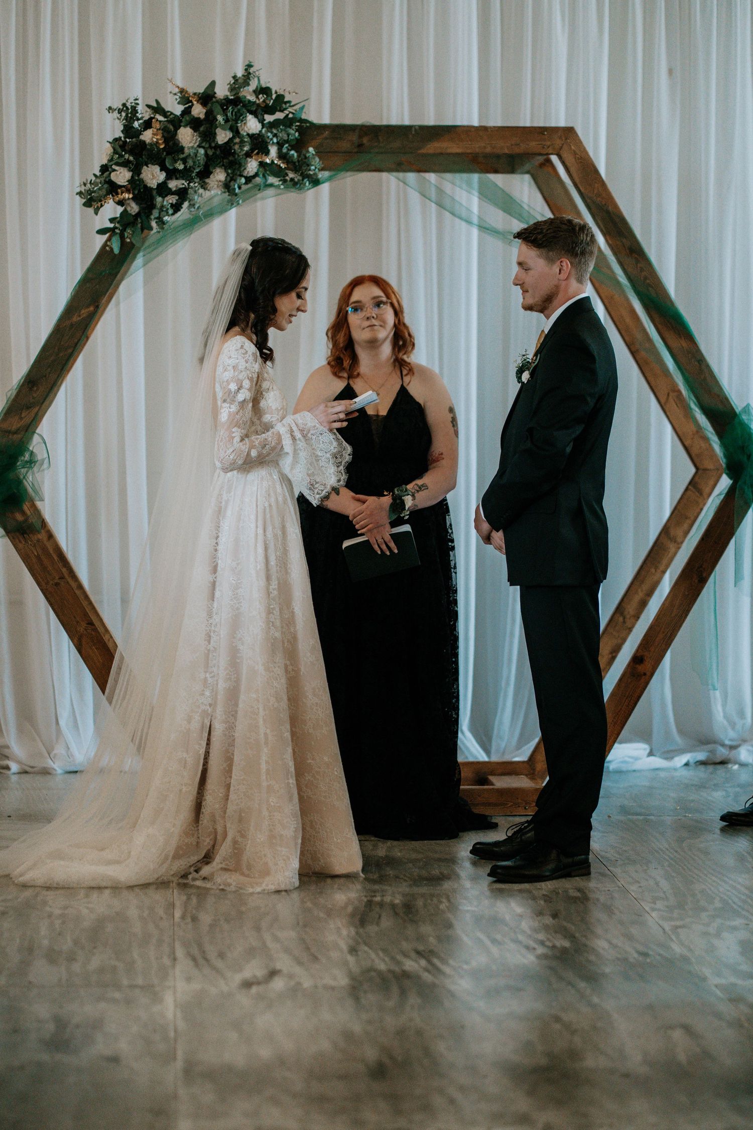 Bride and groom exchange vows under a hexagonal wooden arch with floral decor, officiated by a woman in black.