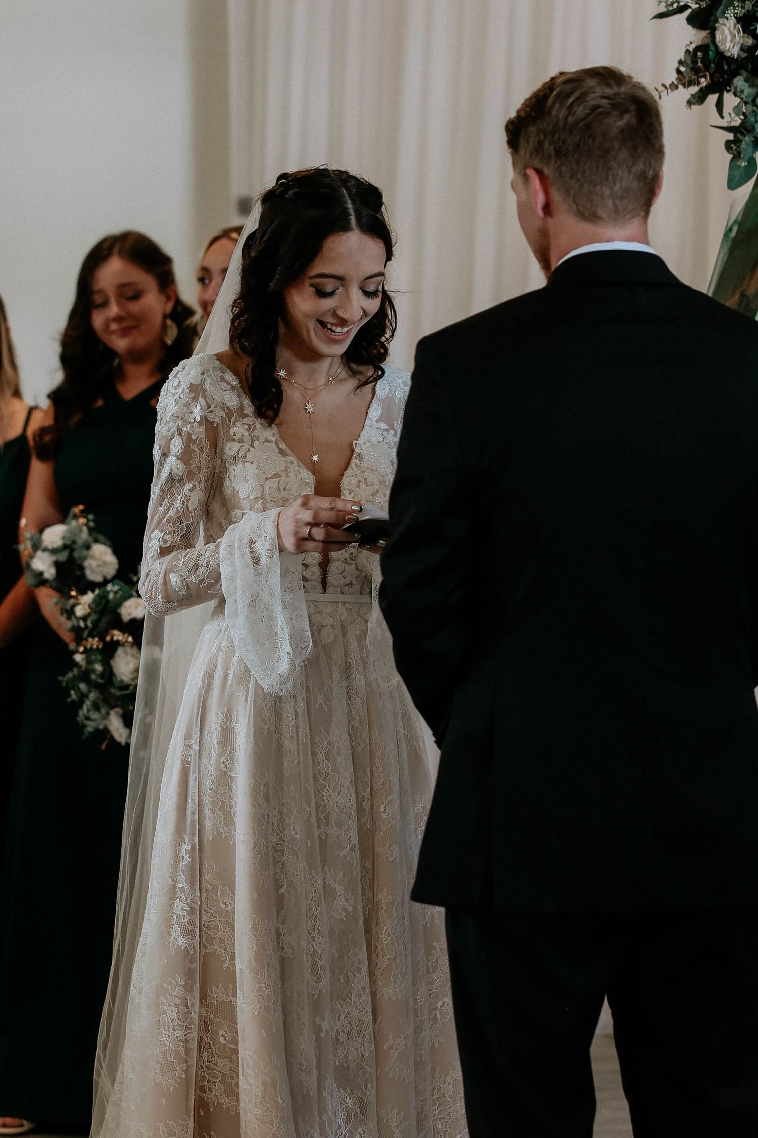Bride in lace long-sleeve wedding gown smiling while exchanging rings with groom during indoor wedding ceremony.