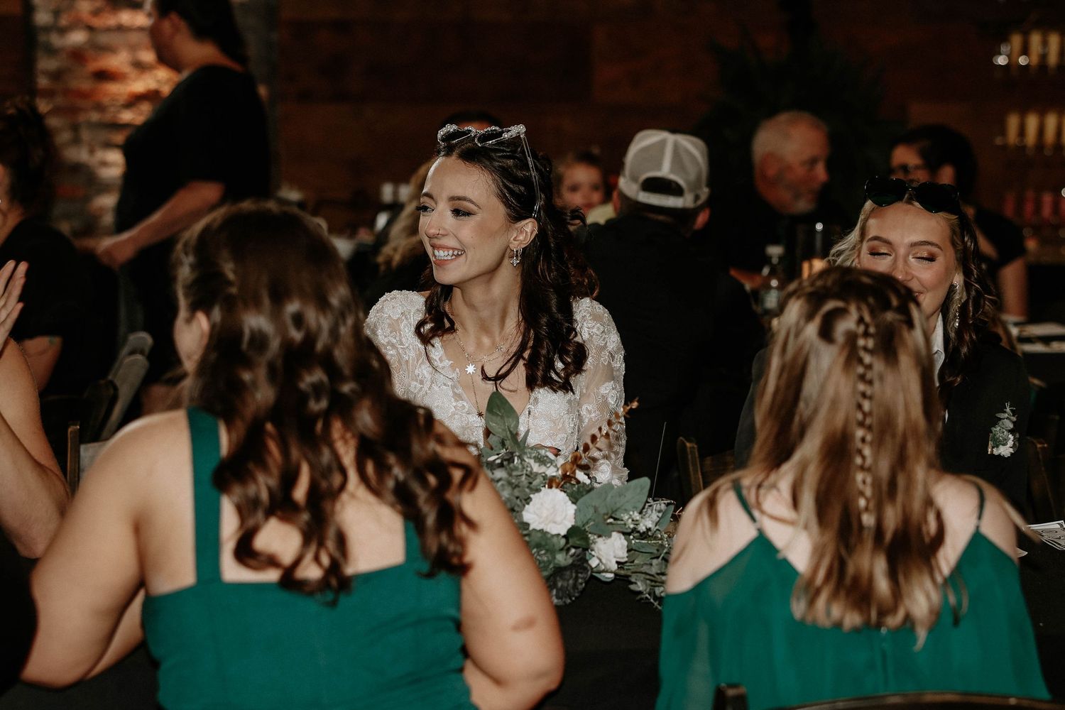 Bride in white lace dress smiles holding white floral bouquet surrounded by bridesmaids in emerald green dresses at wedding reception.