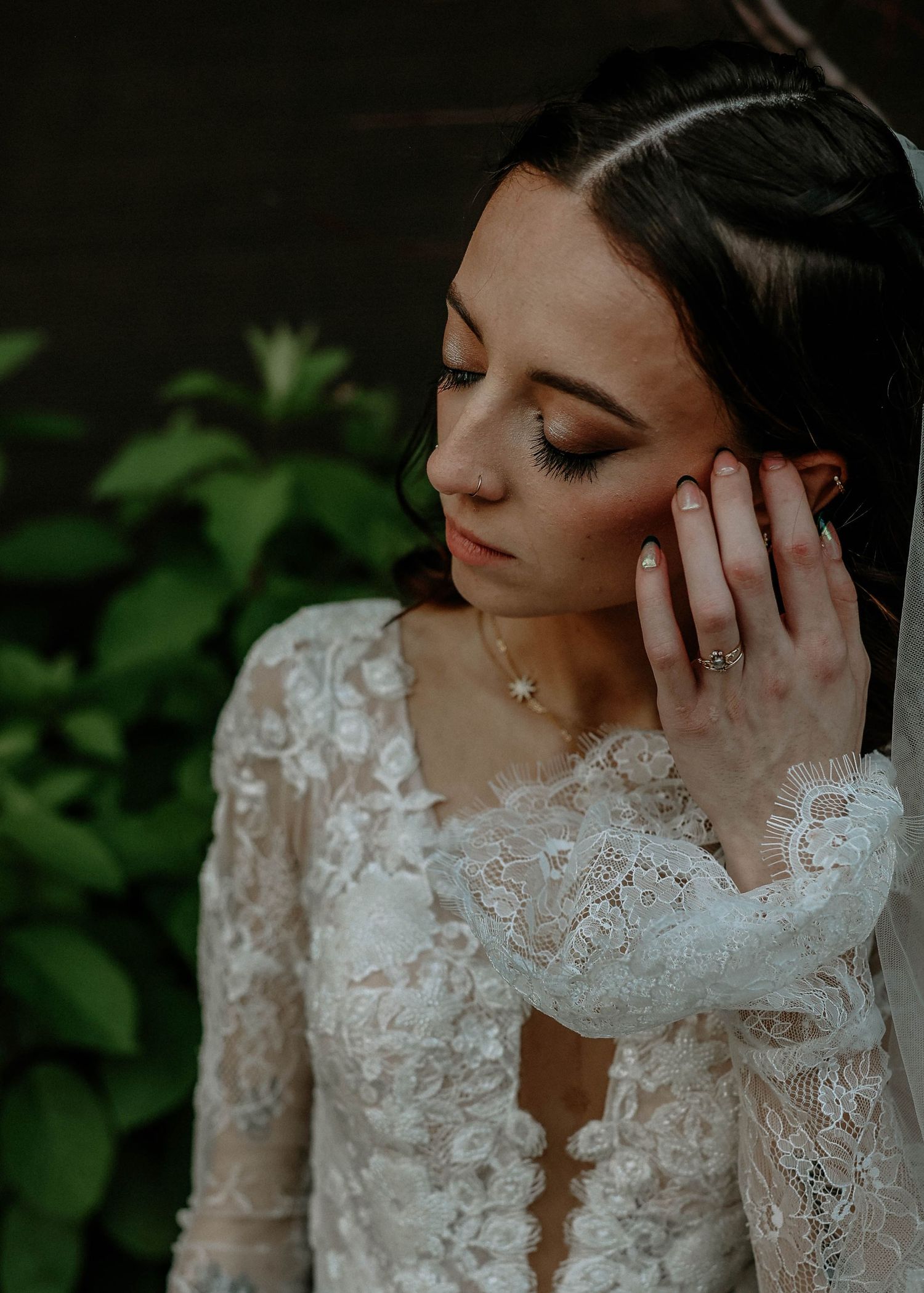 Bride in lace long-sleeve wedding gown touching her ear, wearing a delicate necklace and ring, with green foliage background.