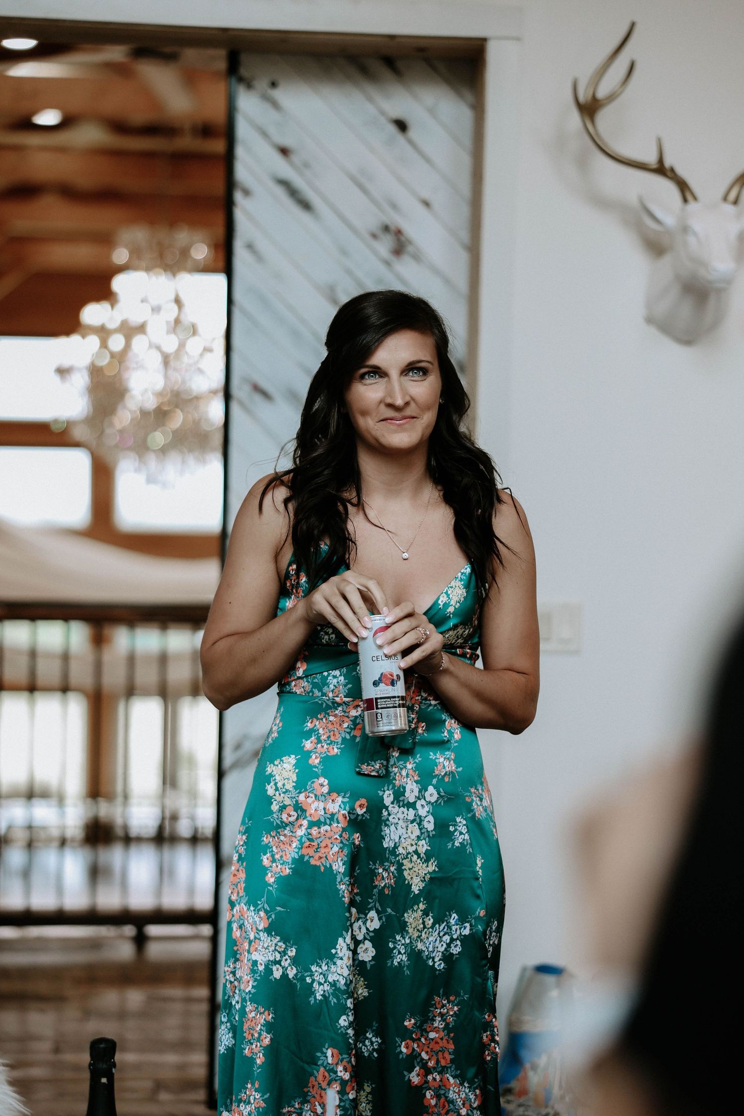 Woman in green floral dress holding small item standing in rustic barn wedding venue interior.
