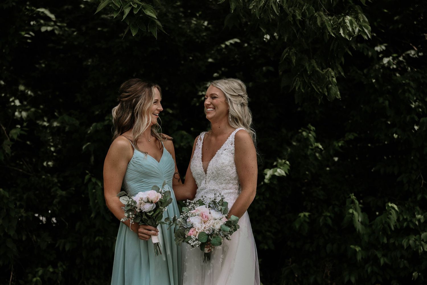 Bride in white lace gown and bridesmaid in mint dress smiling together holding pink bouquets outdoors.