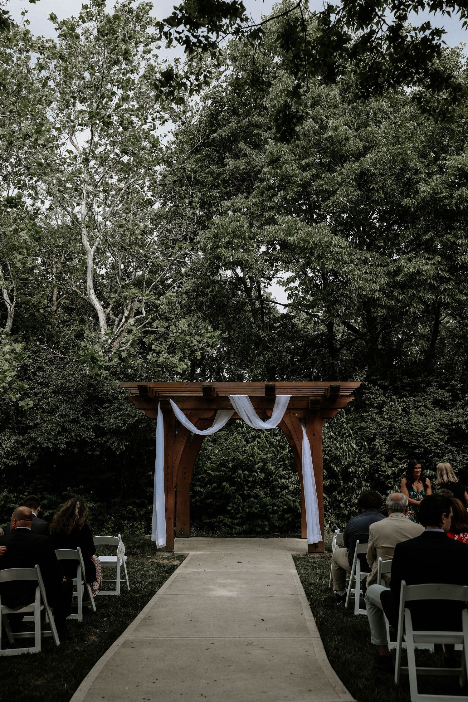 Outdoor wedding ceremony aisle with wooden pergola draped in white fabric, surrounded by lush green trees.