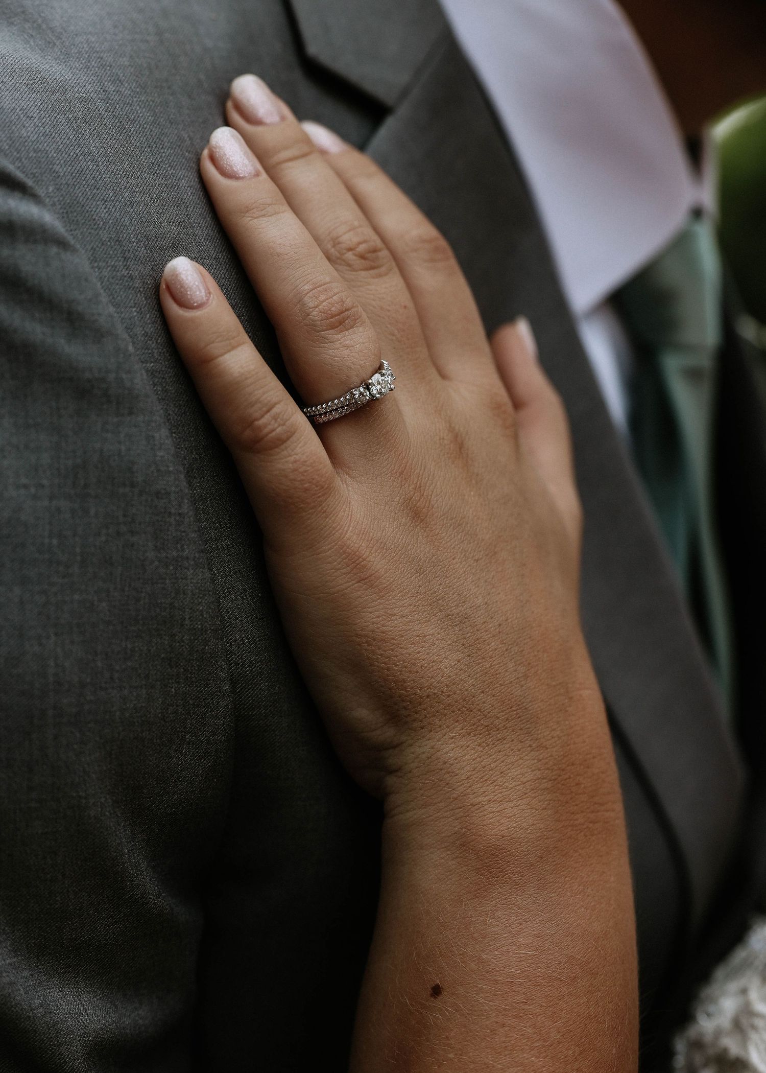 Close-up of a diamond engagement ring on a bride's hand resting on a groom's gray suit.
