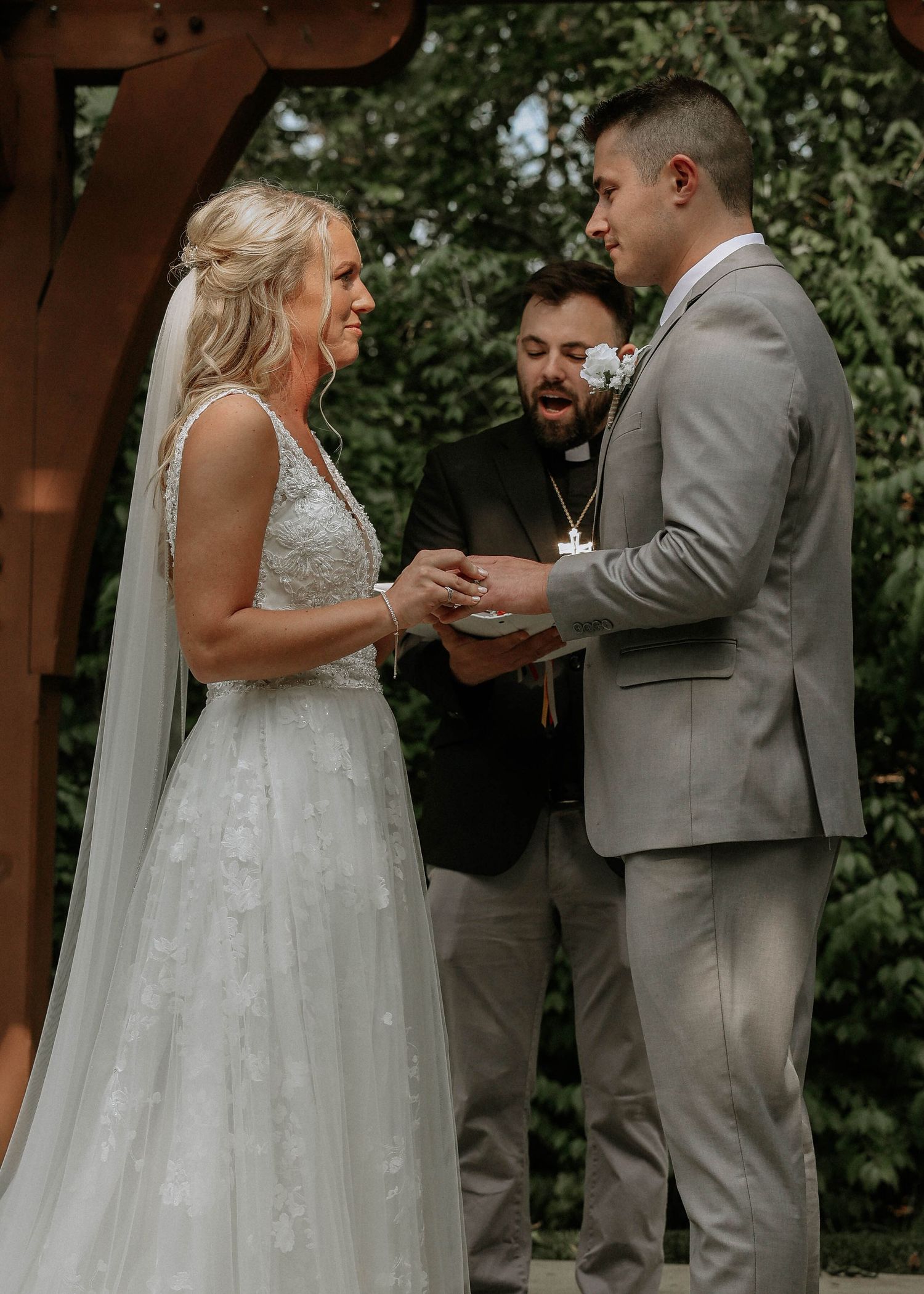 Bride placing wedding ring on groom's finger during outdoor ceremony under wooden arch with officiant.