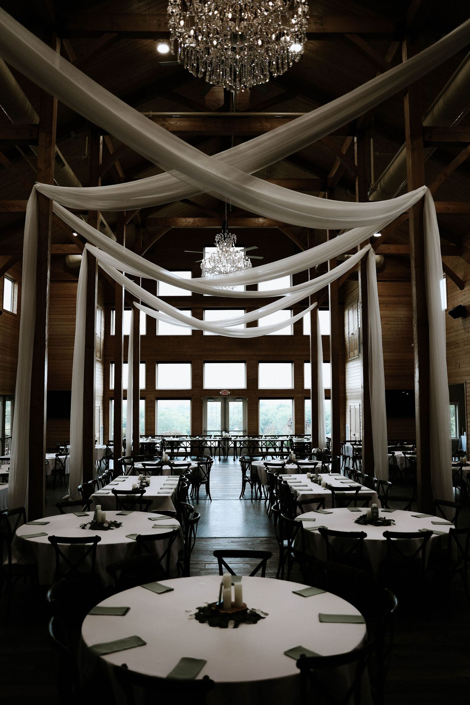 Elegant barn wedding venue with draped white fabric, crystal chandeliers, and round tables set for a reception.