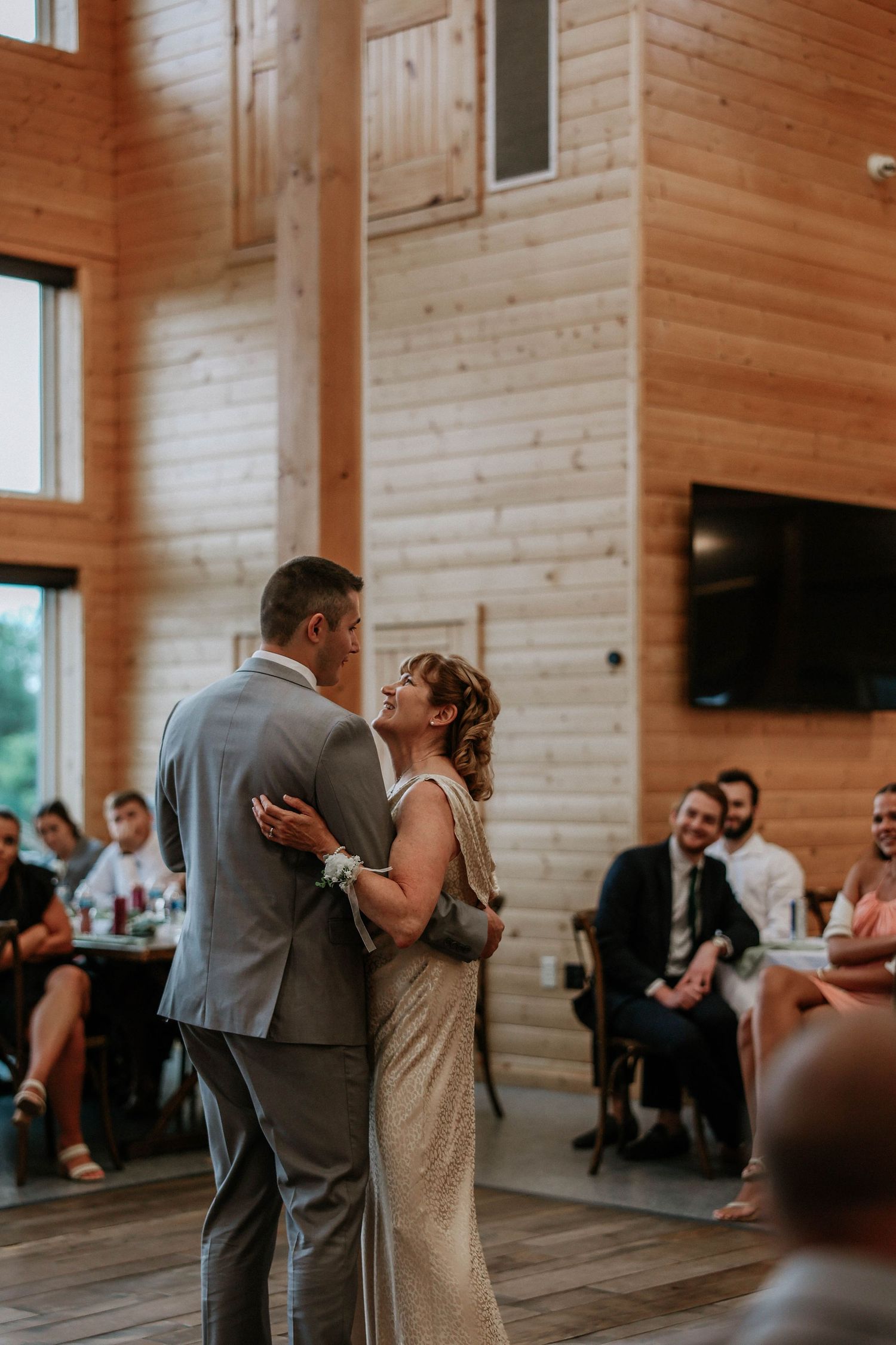 Bride and groom share first dance in rustic wood-paneled wedding venue while guests watch and smile.