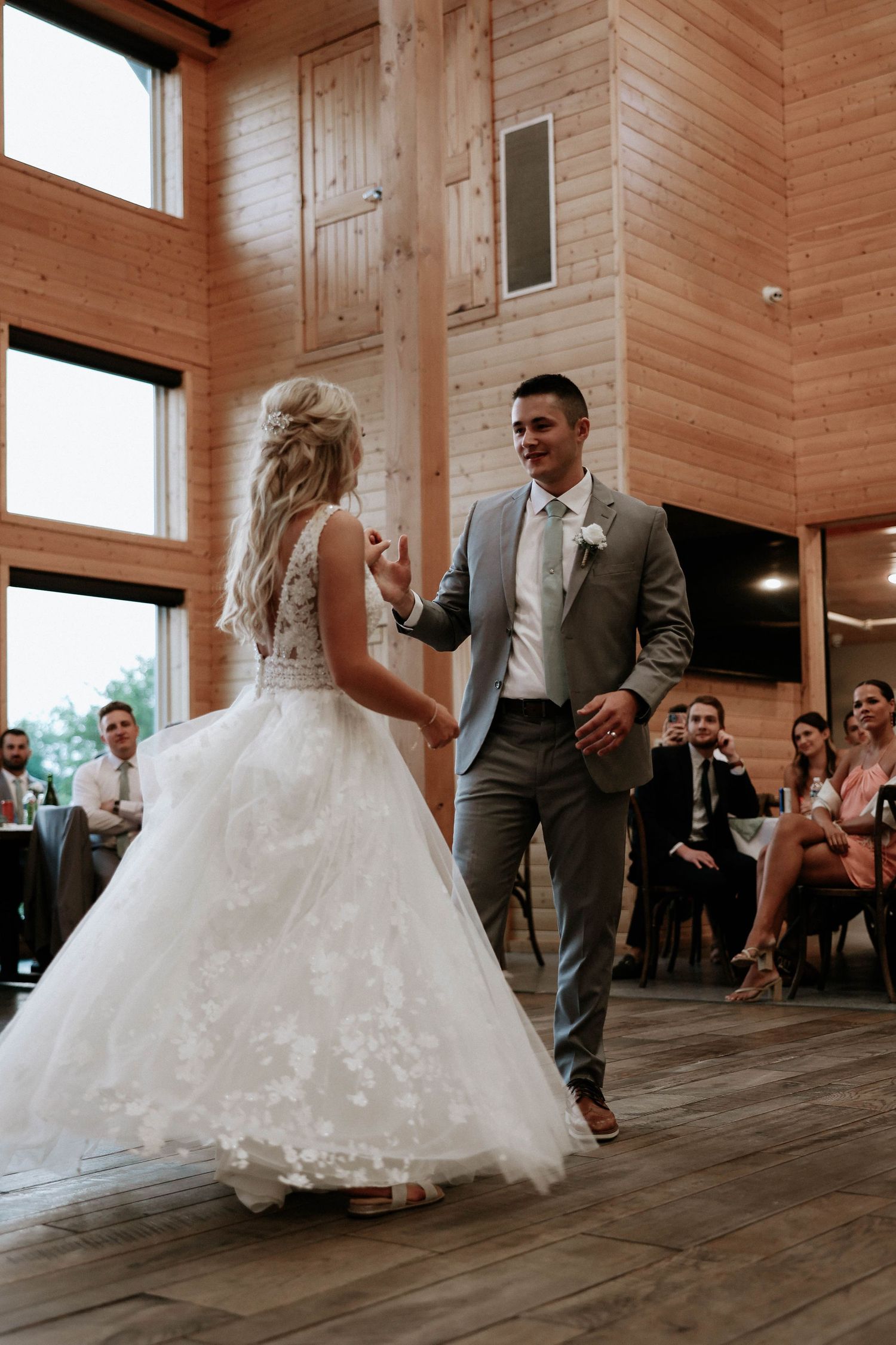 Groom in gray suit smiles at bride in lace wedding gown during first dance in a rustic barn venue.
