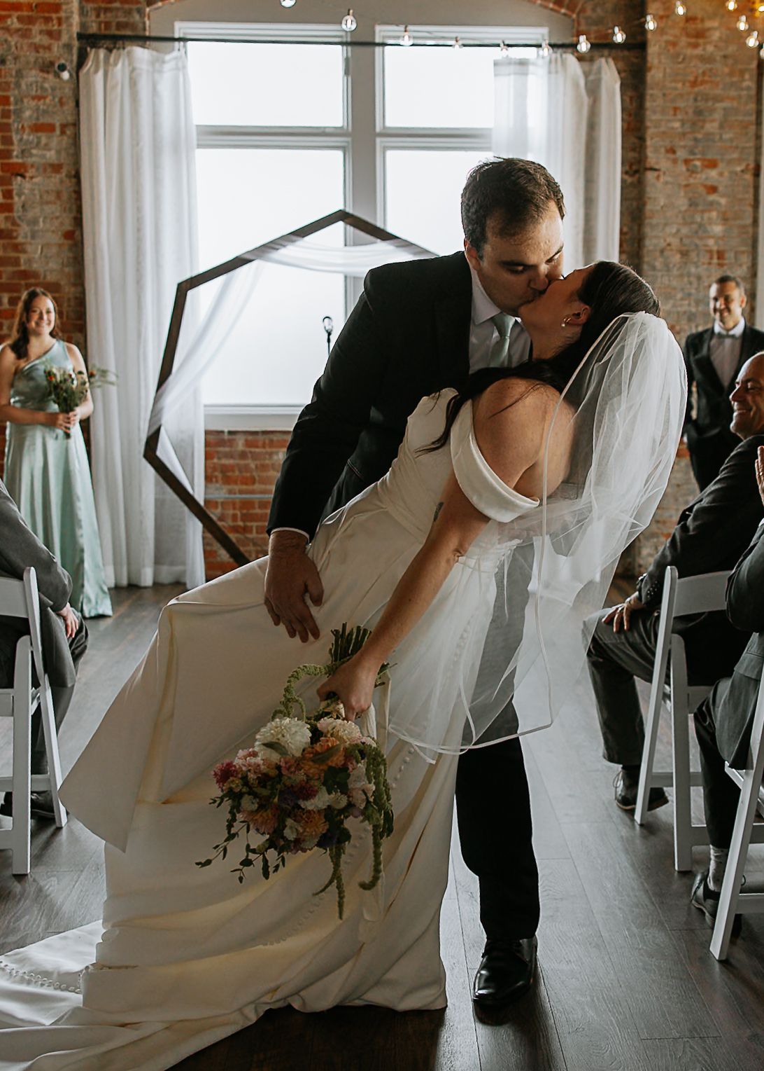 Bride and groom share a romantic dip kiss at the wedding altar with guests seated on either side.