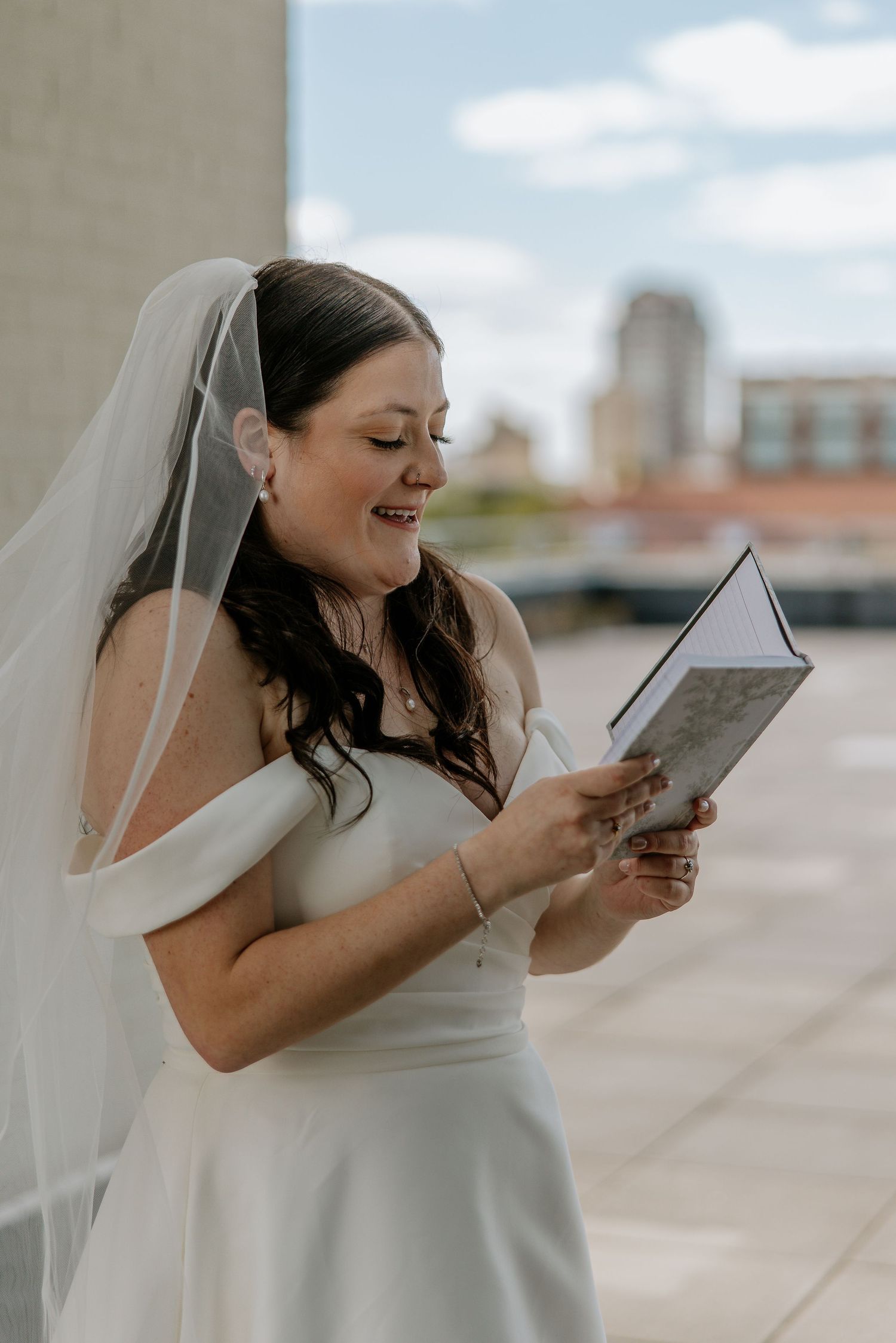 Smiling bride in white gown and veil reads handwritten wedding vows from a book on a rooftop.