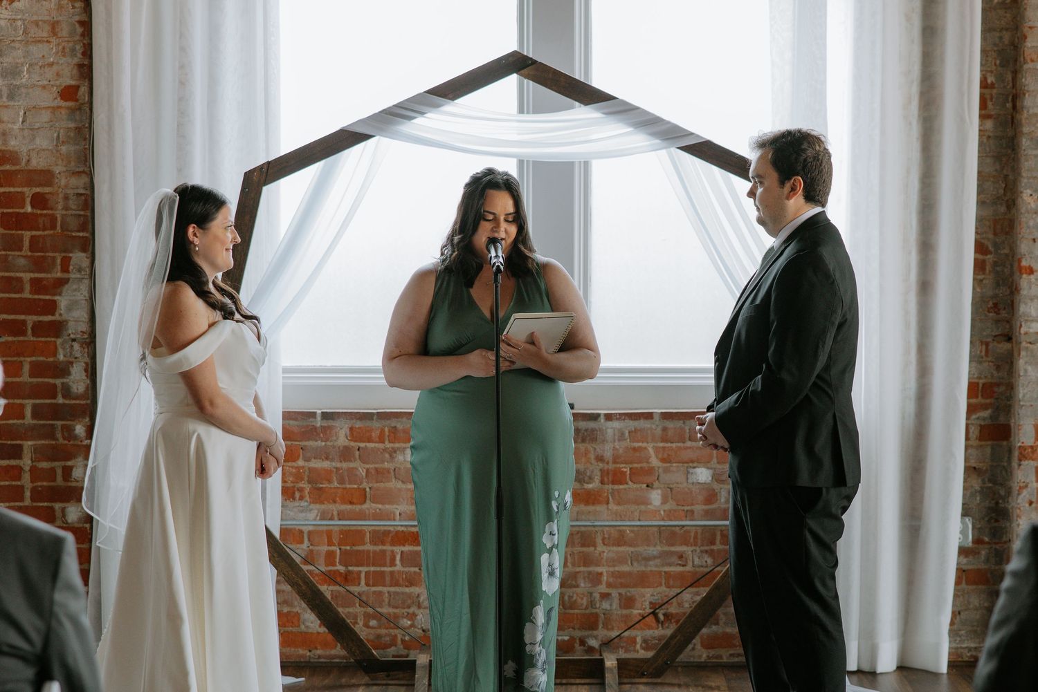 Wedding officiant in green dress reads vows as bride and groom stand at rustic altar.