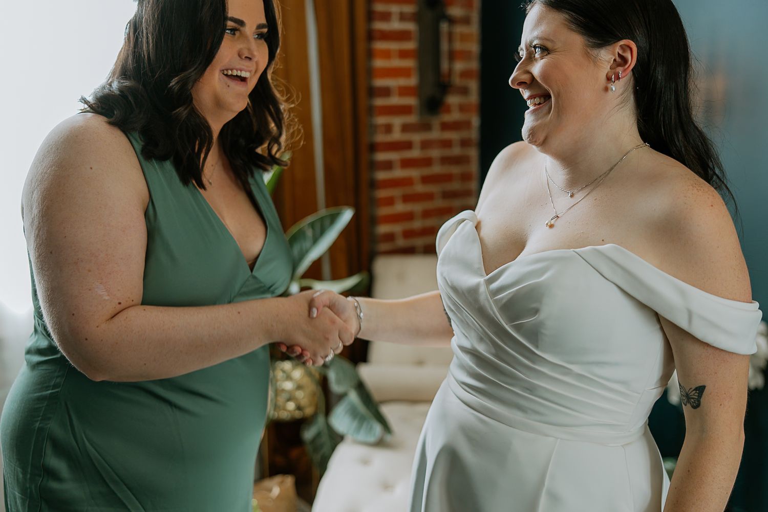 Bride in white off-shoulder gown laughing with bridesmaid in green dress while holding hands.