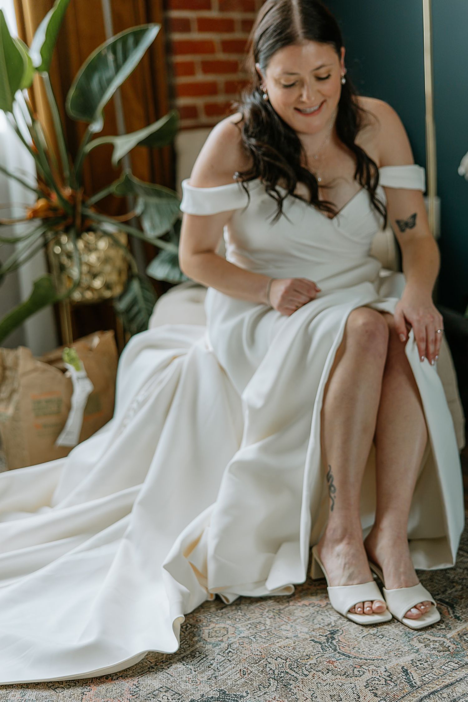 Bride in off-shoulder white gown sitting and adjusting her white heels before wedding.