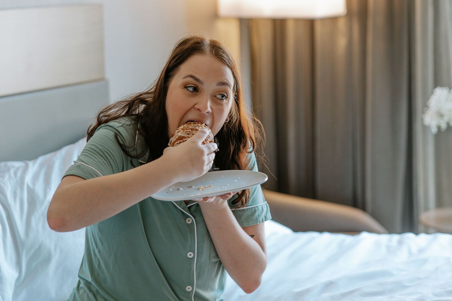 Woman in green pajamas eating a pastry from a white plate while standing in a hotel room.