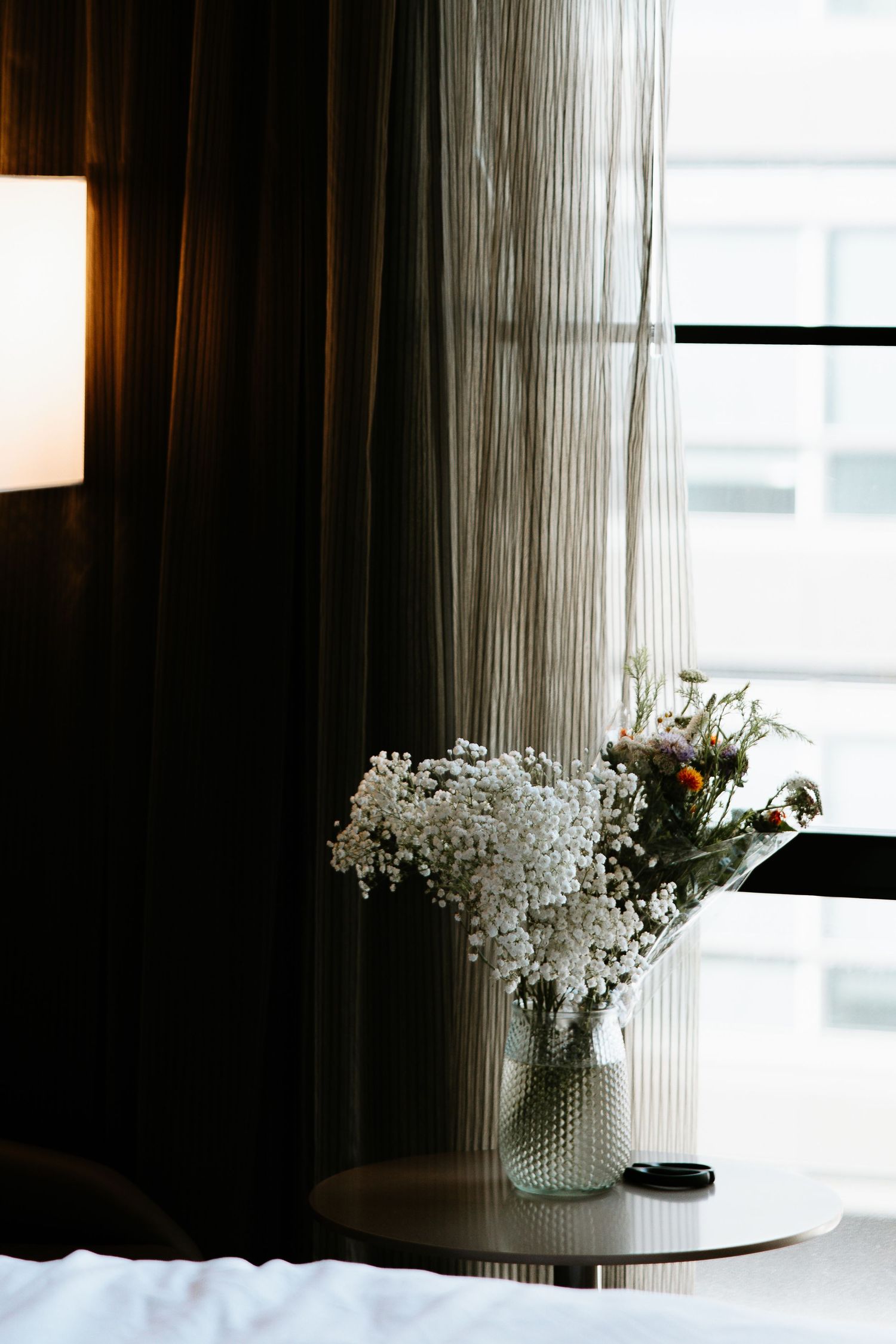 White floral arrangement in a glass vase on a side table beside dark curtains and a window.