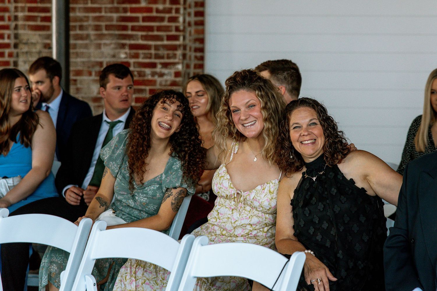 Three smiling women seated together in white chairs at a wedding ceremony.