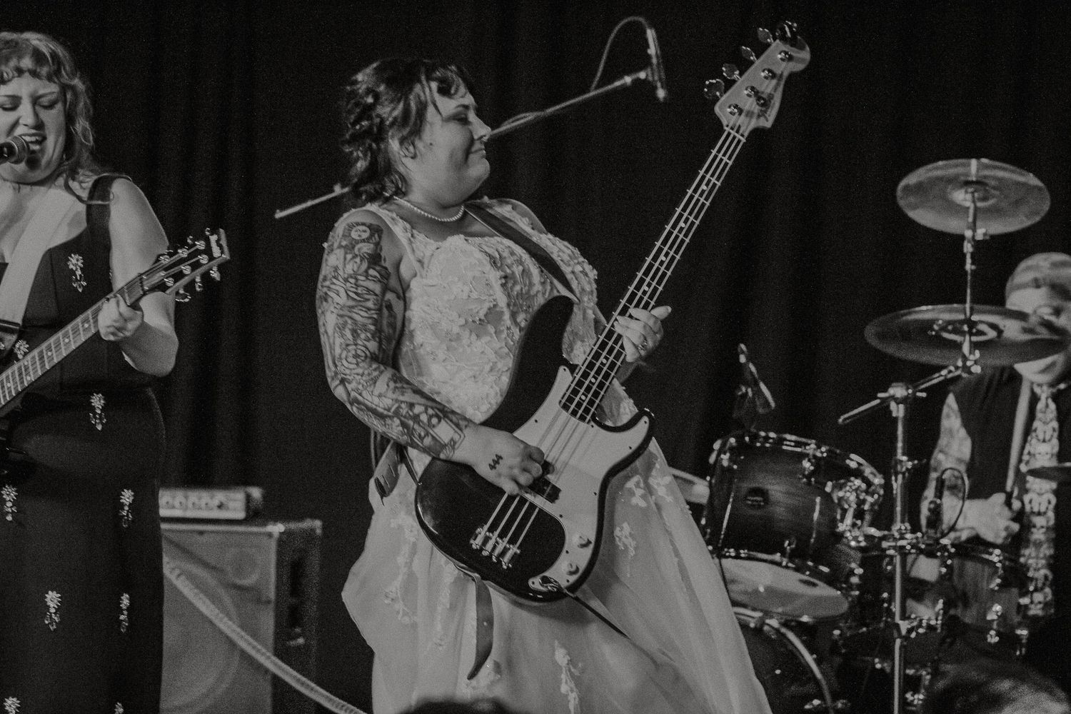 Black and white photo of a bride in a wedding dress playing bass guitar on stage.