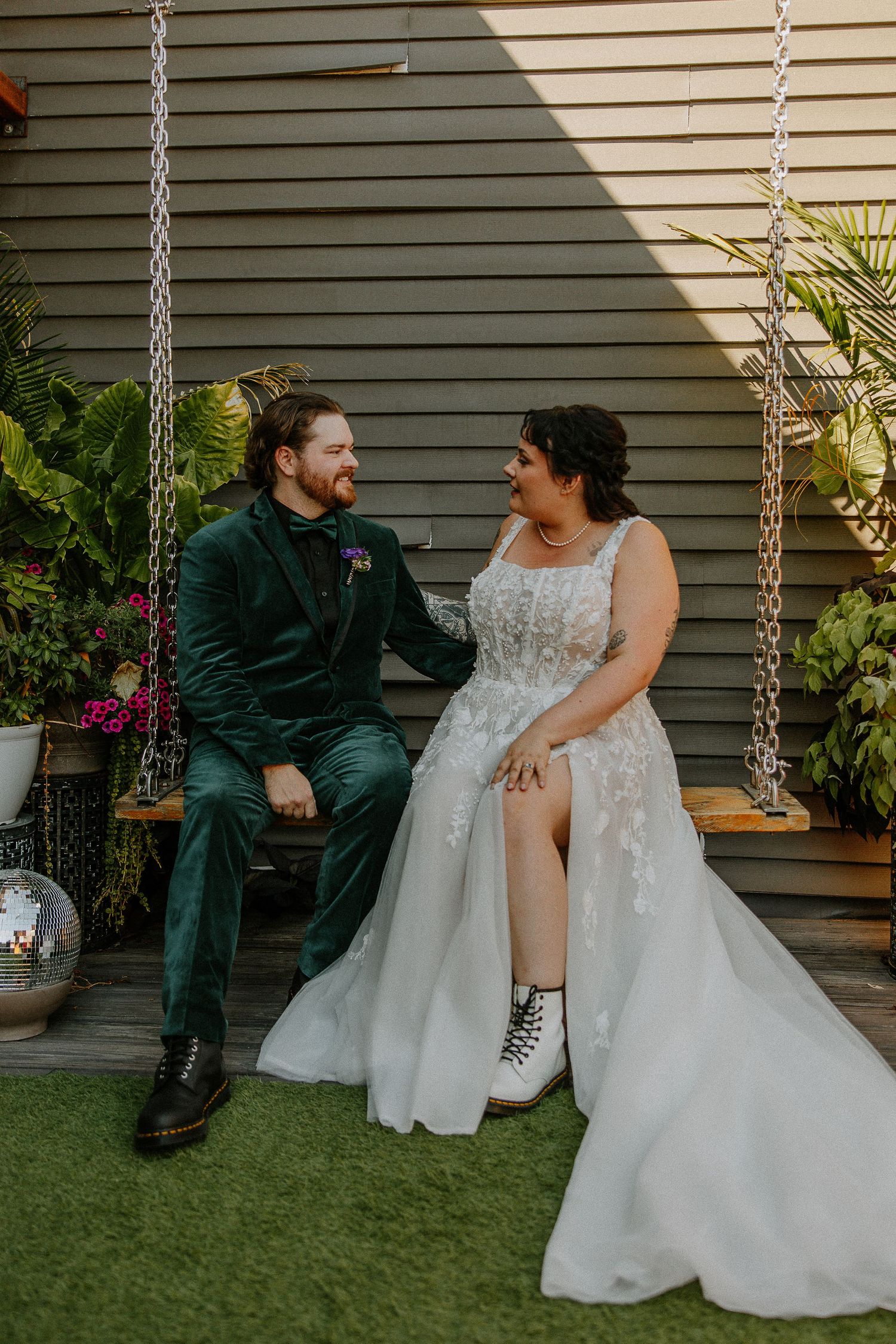Bride in lace gown with combat boots and groom in green velvet suit sit on swing surrounded by plants.