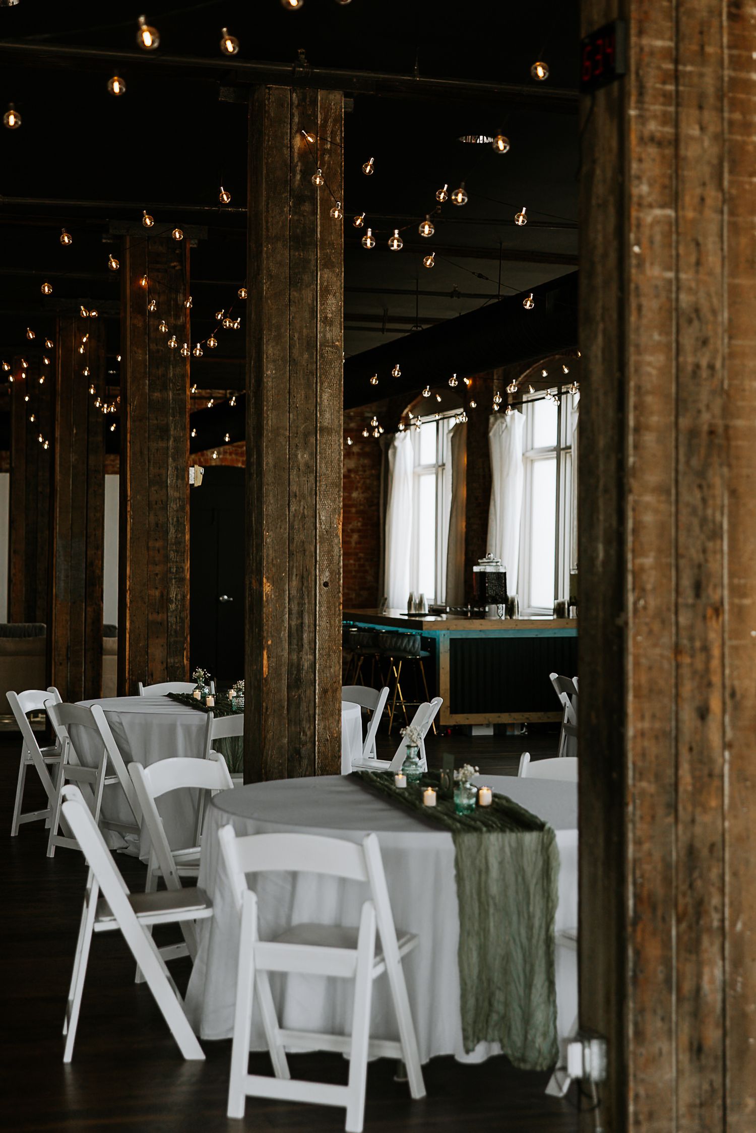 Elegant wedding reception venue with string lights, wooden beams, white chairs, and sage green table runners at The Remedy in Dayton, Ohio