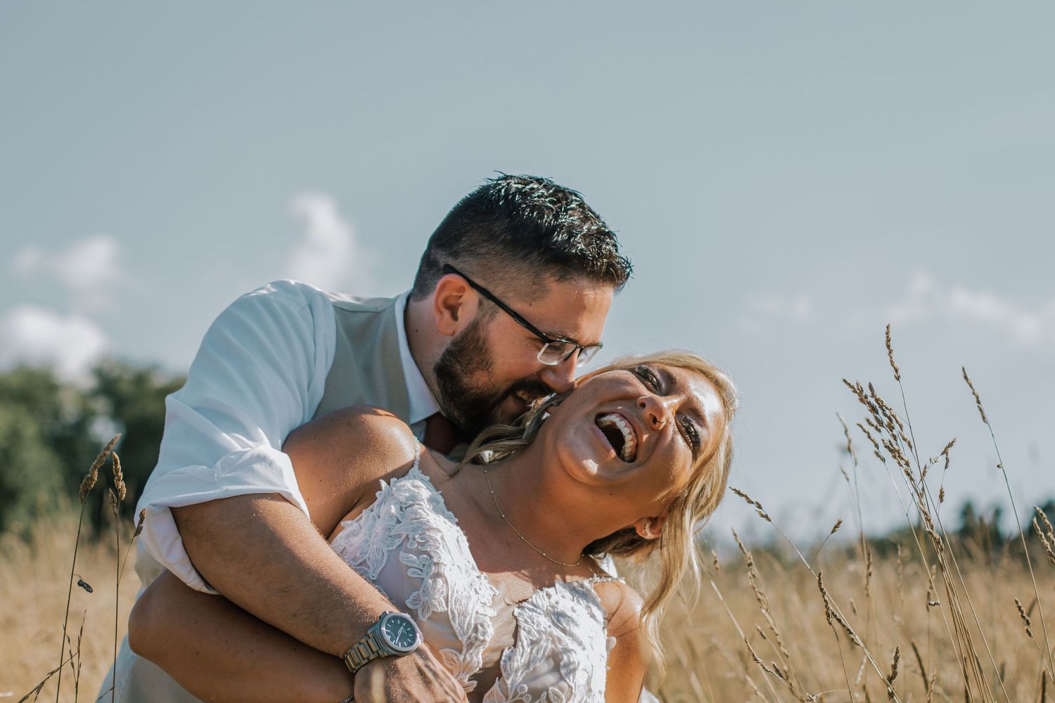 Groom laughing and embracing bride in golden field under blue sky during outdoor engagement session at The Barn at Wolf Creek in Akron, Ohio