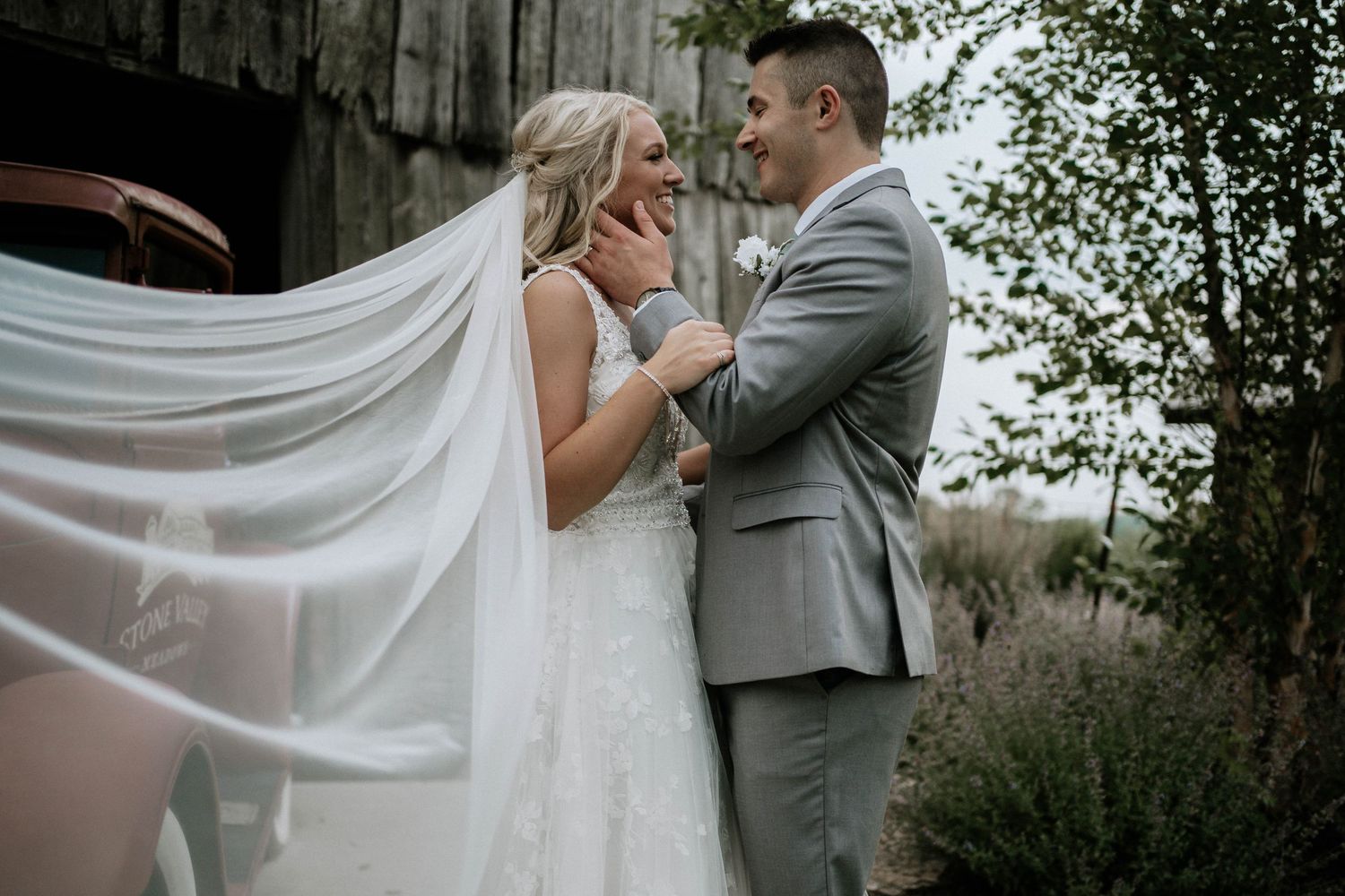 Bride and groom smiling at each other as her veil blows in the wind near a rustic barn.