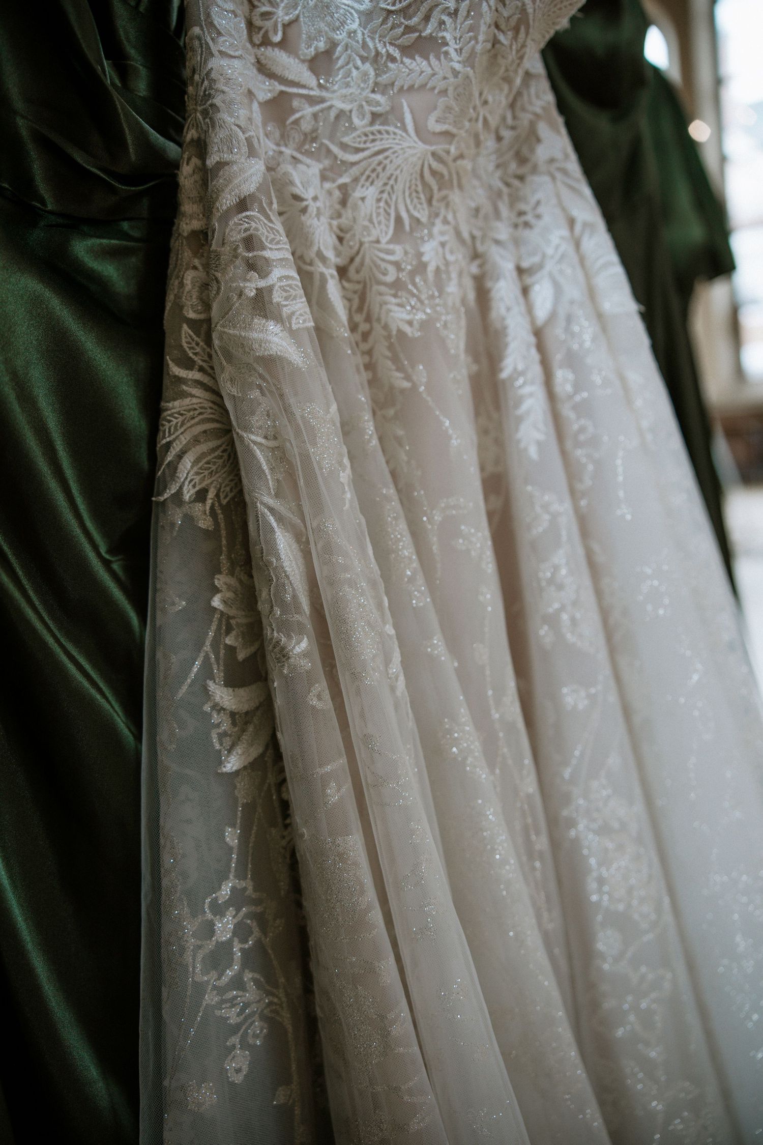 Close-up of intricate lace detailing on a white wedding gown hanging beside emerald bridesmaid dresses at Urban Artifact in Cincinnati, Ohio
