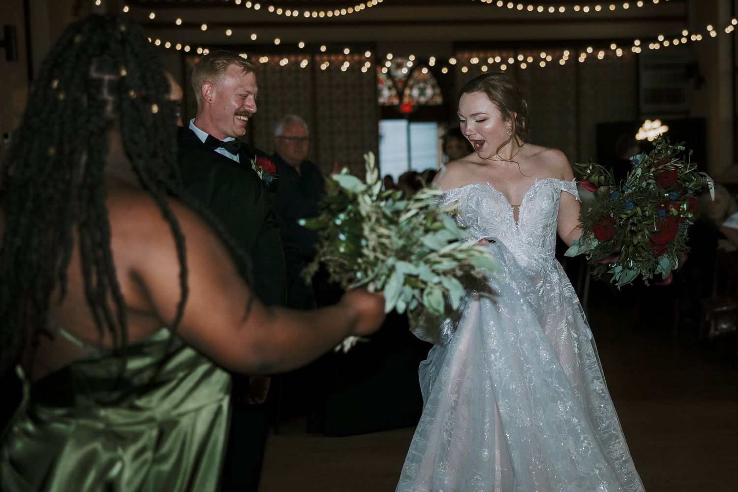Bride in lace gown receives bouquet from guest at elegant wedding reception with string lights at Urban Artifact in Cincinnati, Ohio