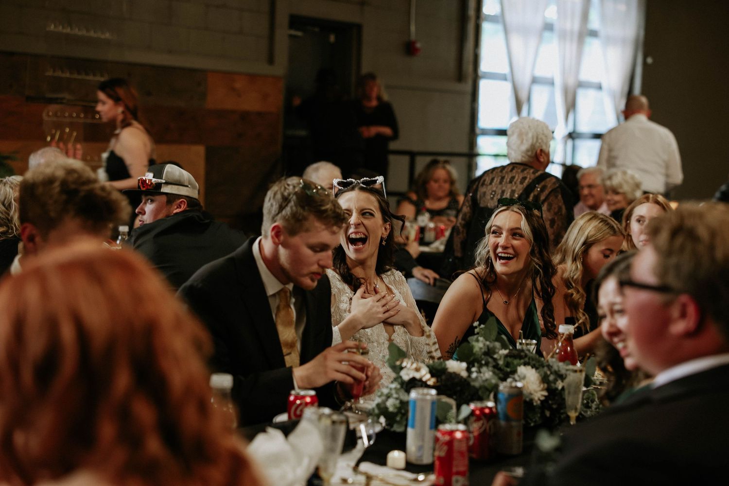 Guests laughing and celebrating at a wedding reception dinner table in a rustic venue.
