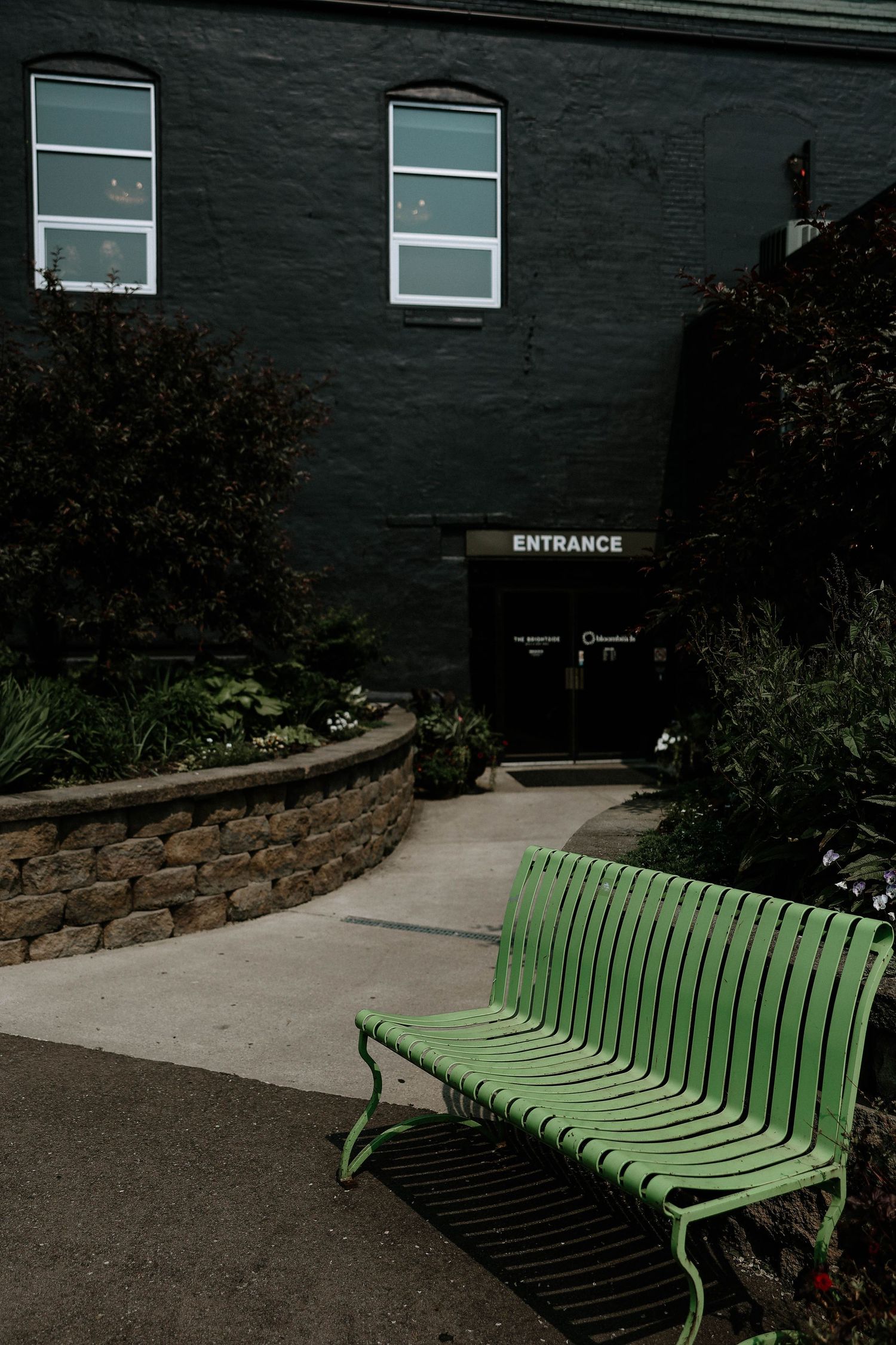 Green metal bench outside a dark brick building entrance, surrounded by lush garden plants and a stone retaining wall.
