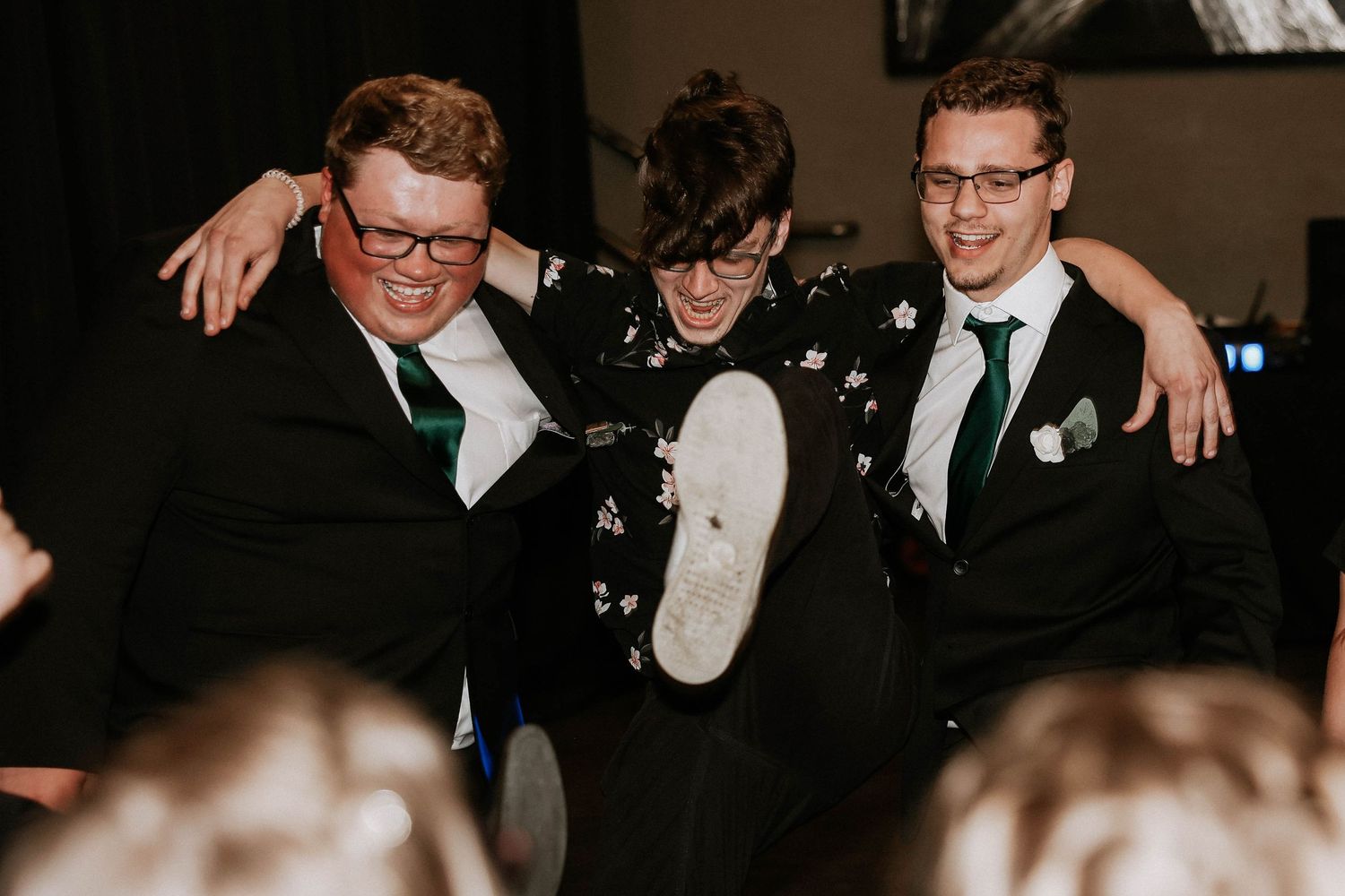 Three men in suits celebrating at a formal event, laughing and kicking up their heels together.