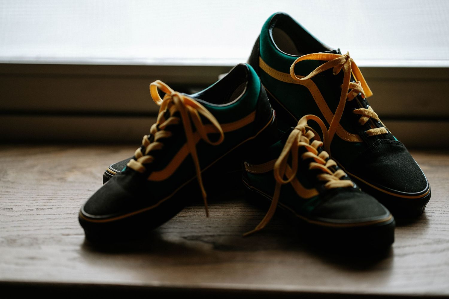 Dark green and yellow Vans sneakers with gold laces displayed on a wooden surface near a window.