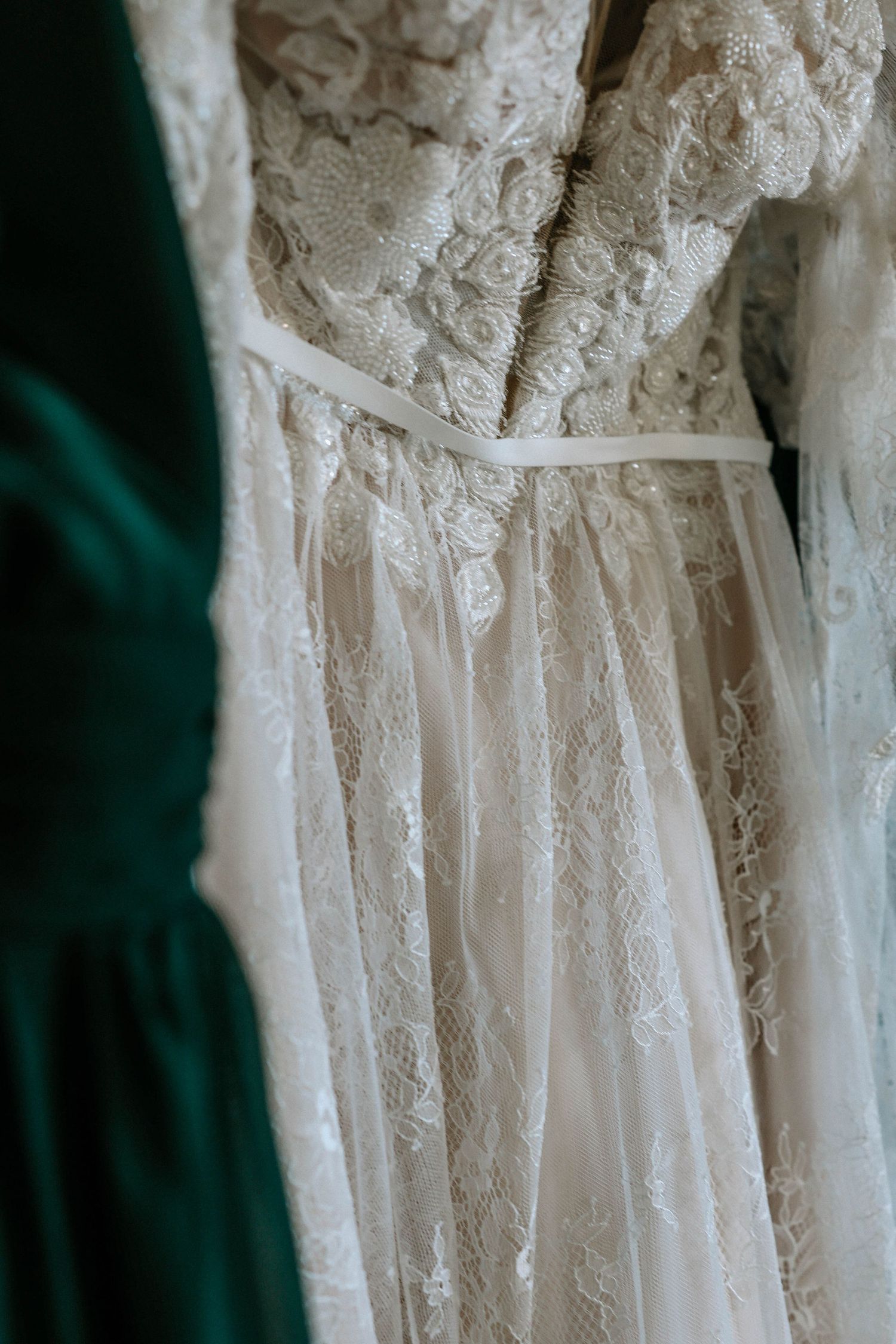 Elegant white lace wedding dress with floral details and ribbon belt hanging on display.