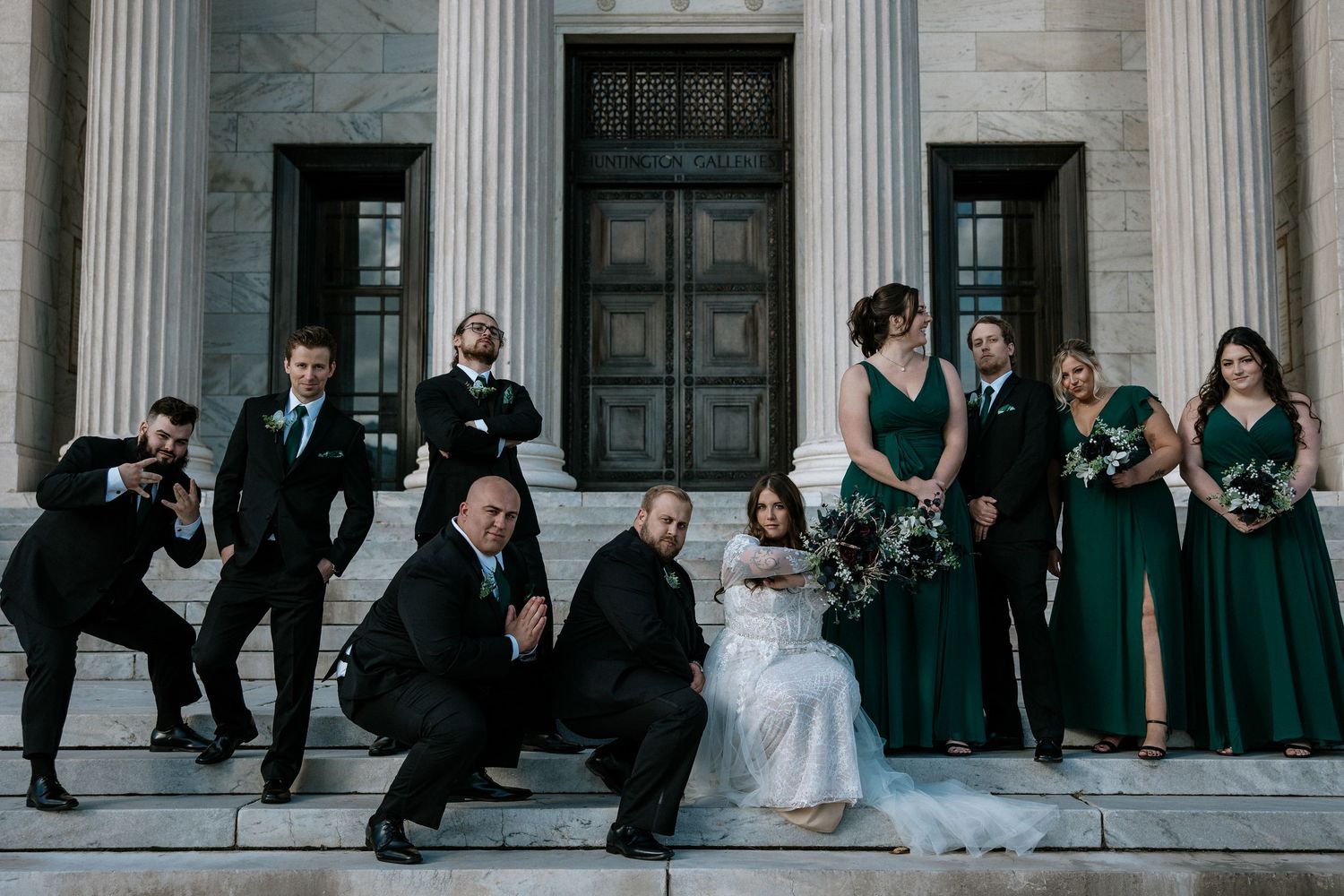 Wedding party strikes fun poses on grand stone steps with columns, bridesmaids in emerald green dresses.