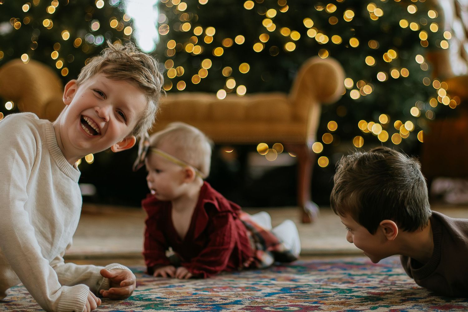 Three children laughing and playing together on a rug in front of a Christmas tree at Mill Street Studios in Cincinnati, Ohio.