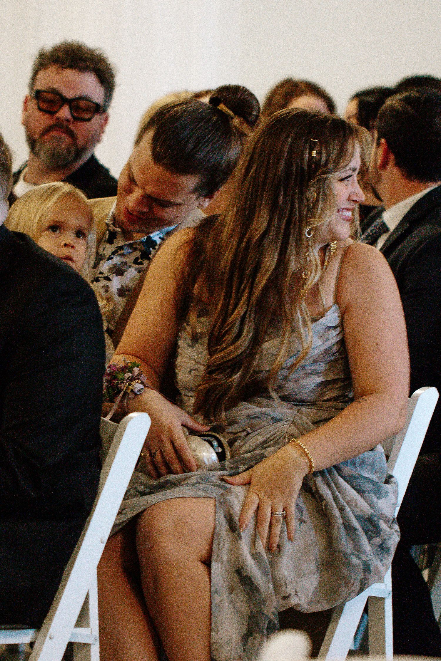Two women laughing together with young child at wedding ceremony seated on white folding chairs.