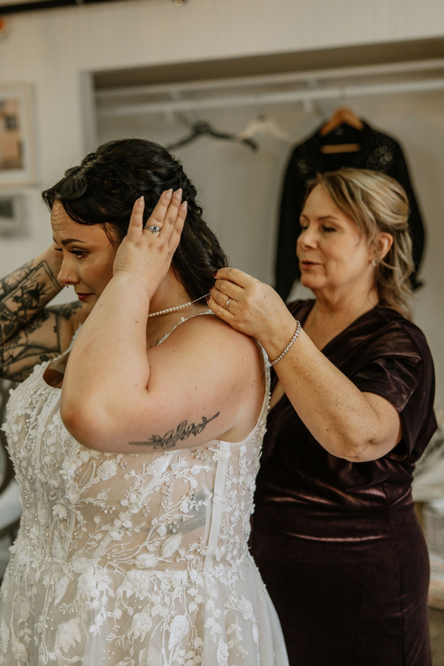Mother fastening a pearl necklace on tattooed bride in white lace wedding dress.