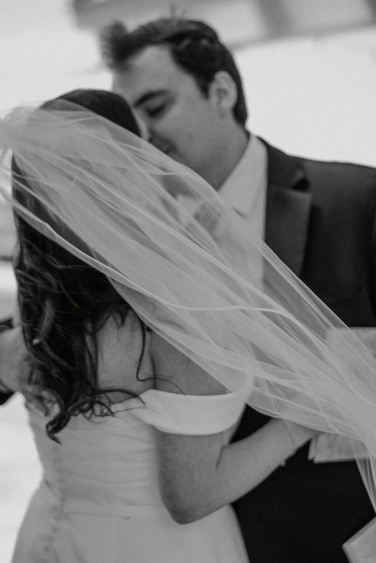 Black and white photo of groom kissing bride as her veil flows during romantic wedding moment.