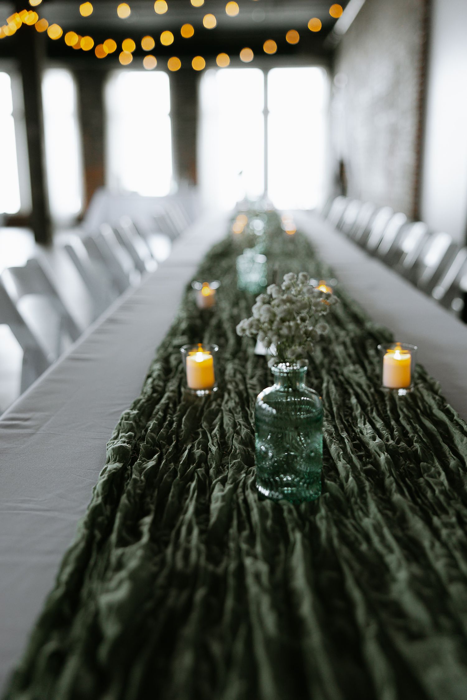 Long wedding reception table with green textured runner, candles, and glass bud vases.