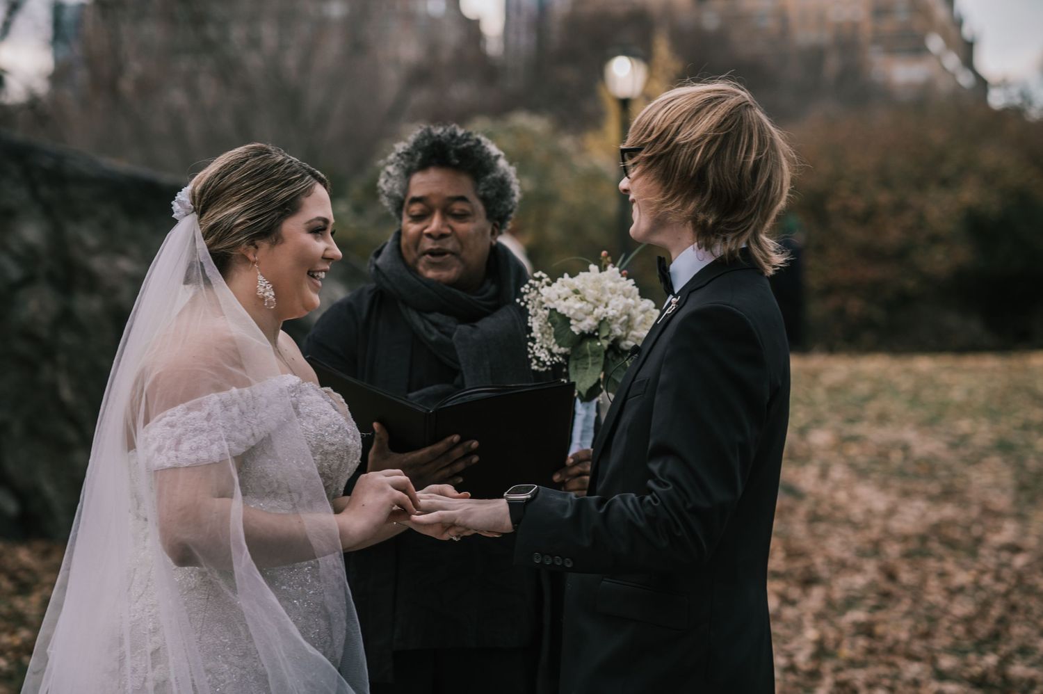 Bride and groom exchanging rings with smiling officiant during intimate Central Park wedding ceremony.