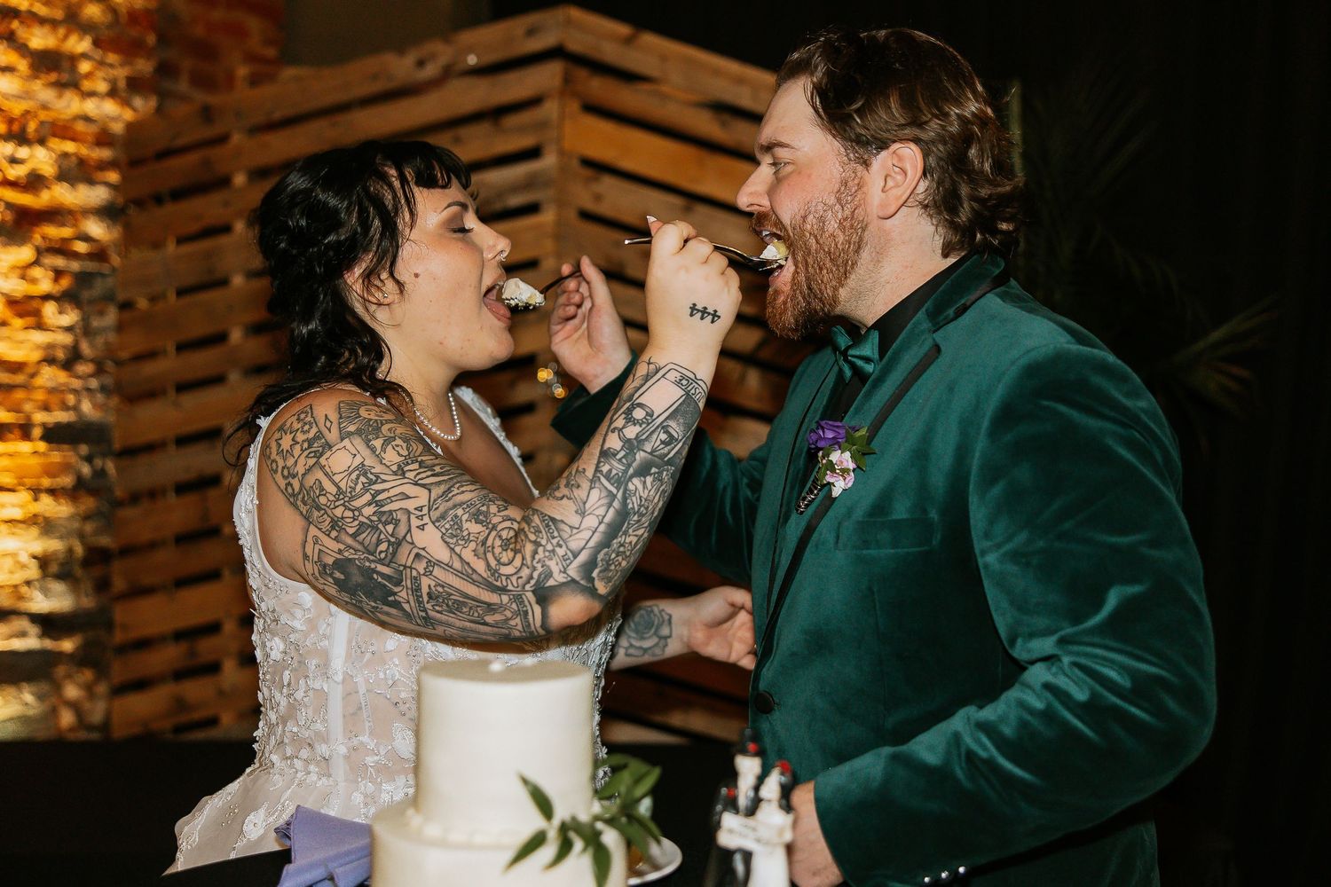 Bride with tattoos feeding wedding cake to smiling groom in green velvet suit.