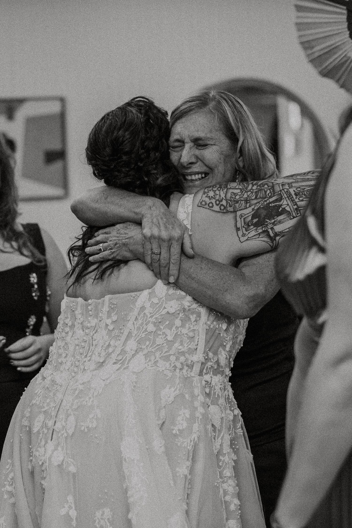 Black and white photo of two women sharing an emotional hug at a wedding, one wearing a lace bridal gown.