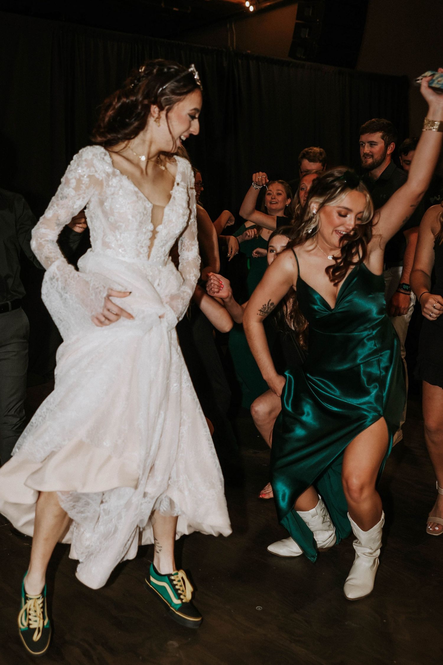 Bride in lace gown and Vans dances with guest in emerald satin dress at lively wedding reception.