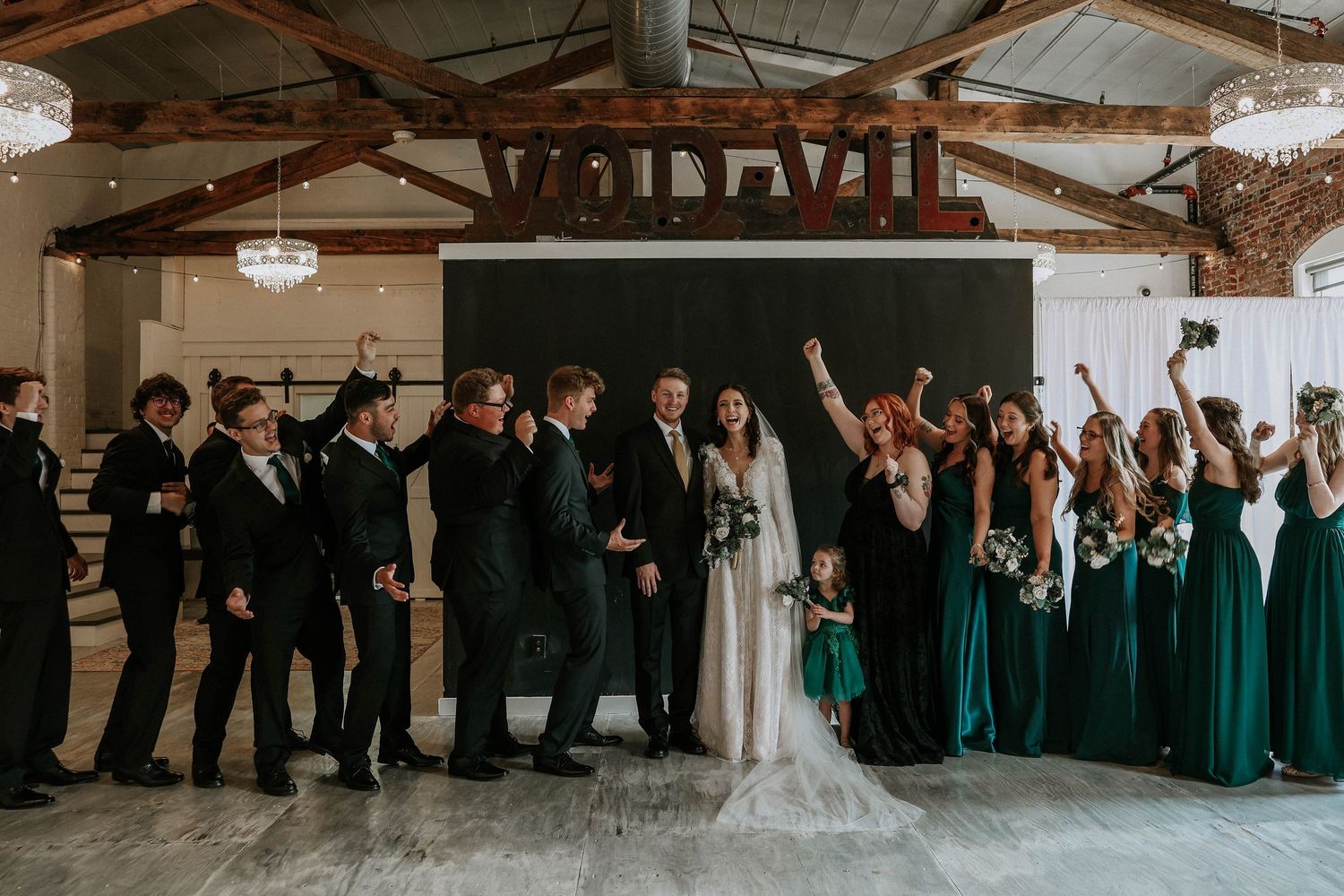 Wedding party celebrates in elegant venue, groomsmen in black suits and bridesmaids in emerald green gowns cheering together.