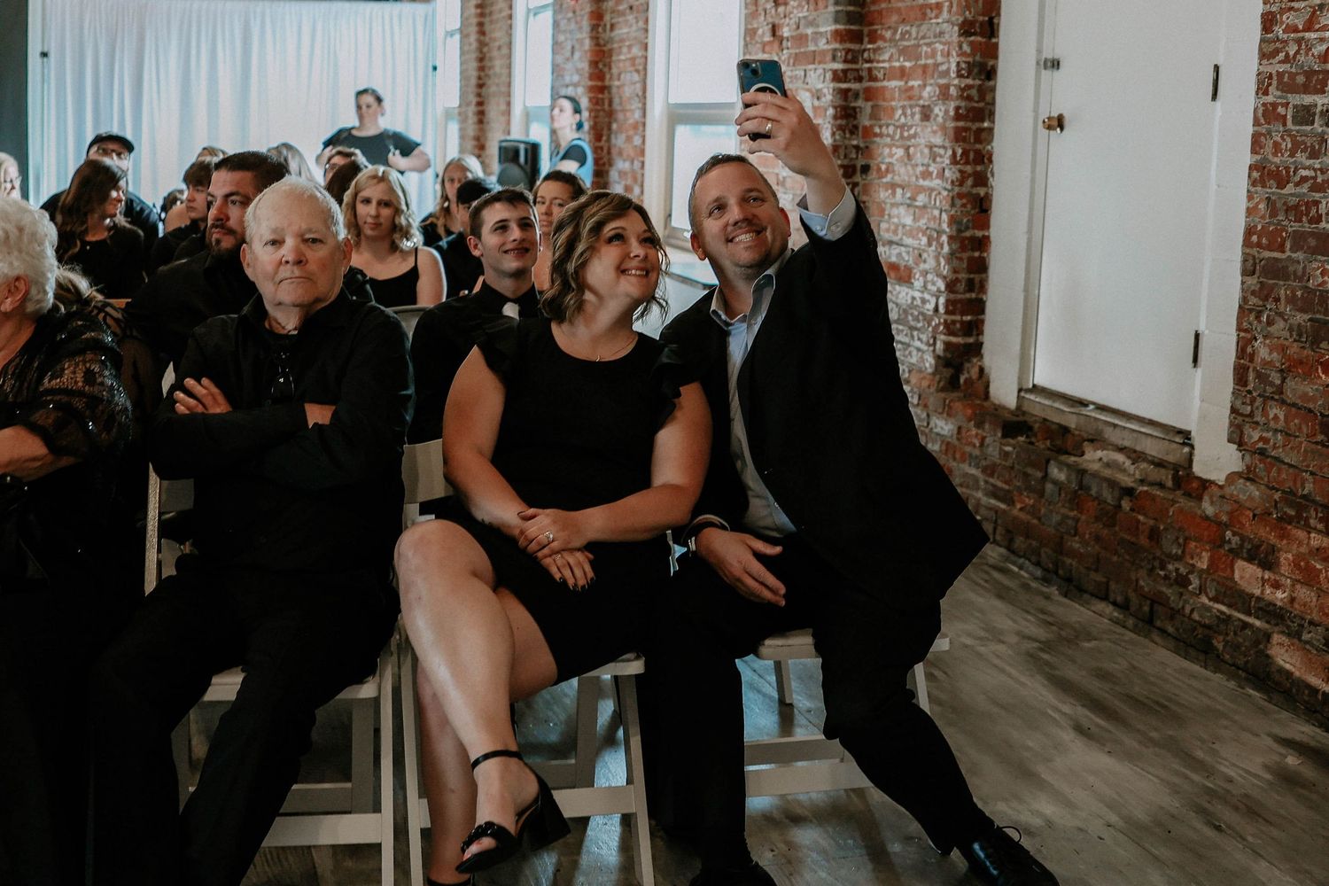 Group of wedding guests smiling and taking a selfie while seated at an indoor ceremony venue.