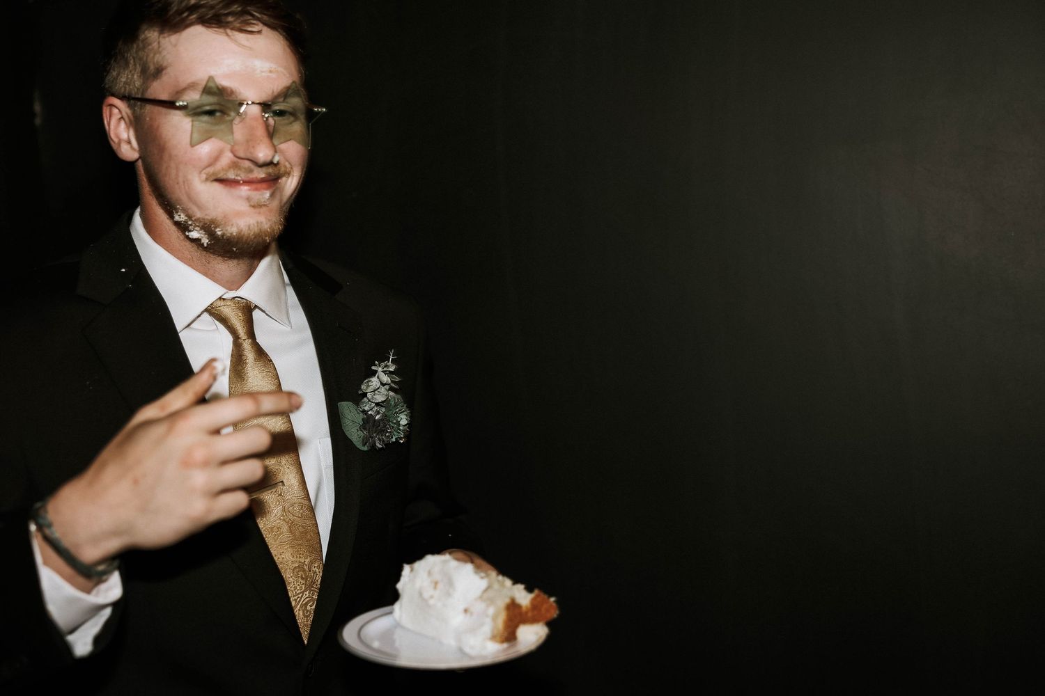 Groom in black suit and gold tie smiling while holding a plate of wedding cake at reception.