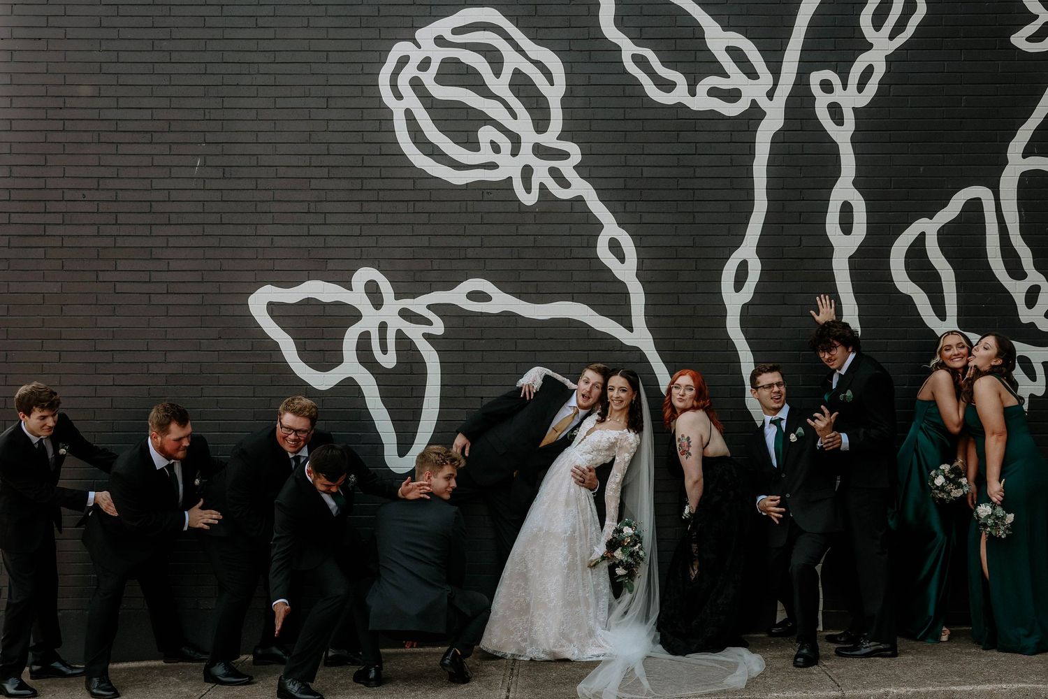 Wedding party poses playfully before a black wall with white floral mural, bride in lace gown holding bouquet.